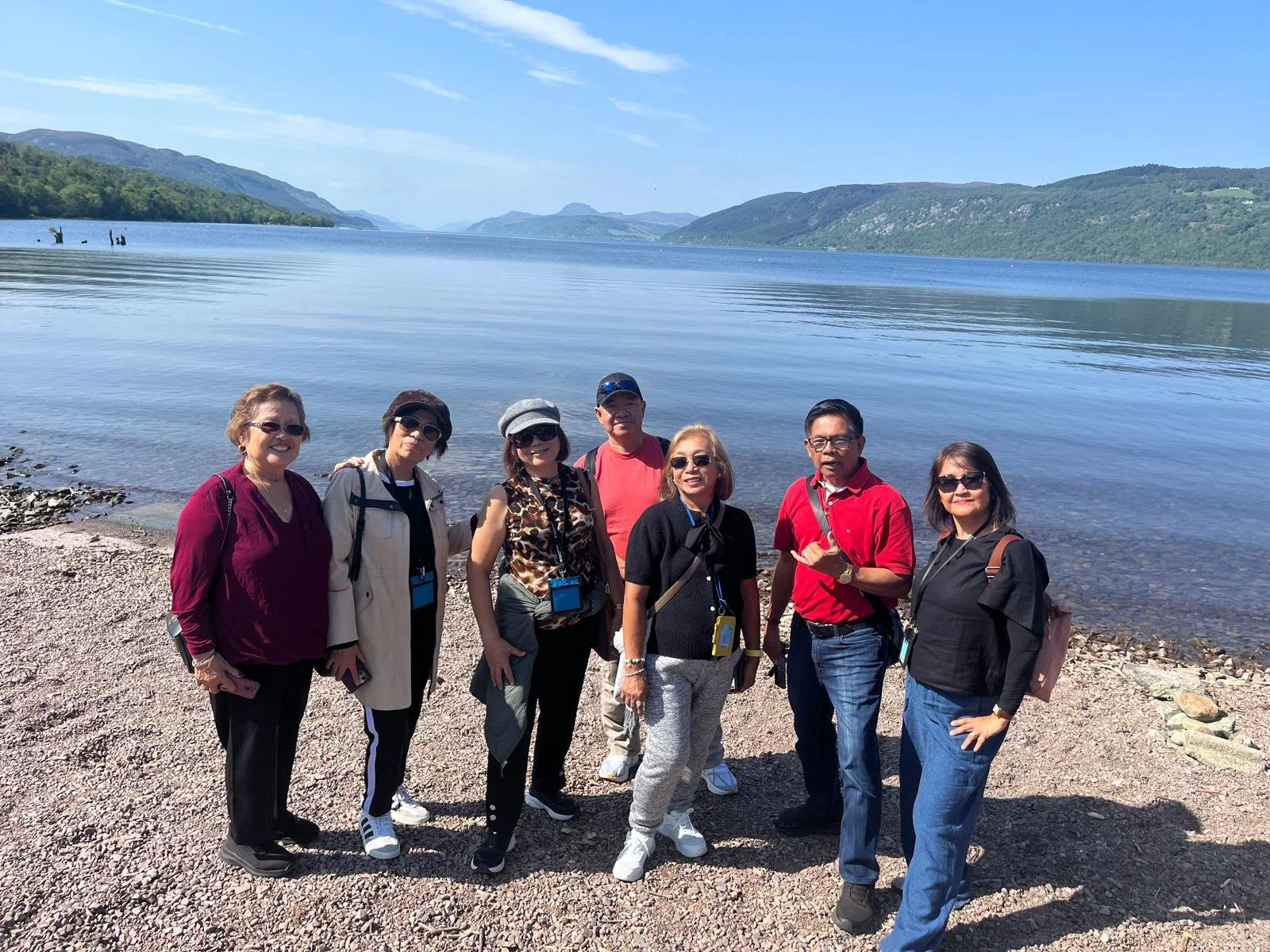 A group of seven people standing on a pebble beach next to a calm lake, with mountains and a blue sky in the background.