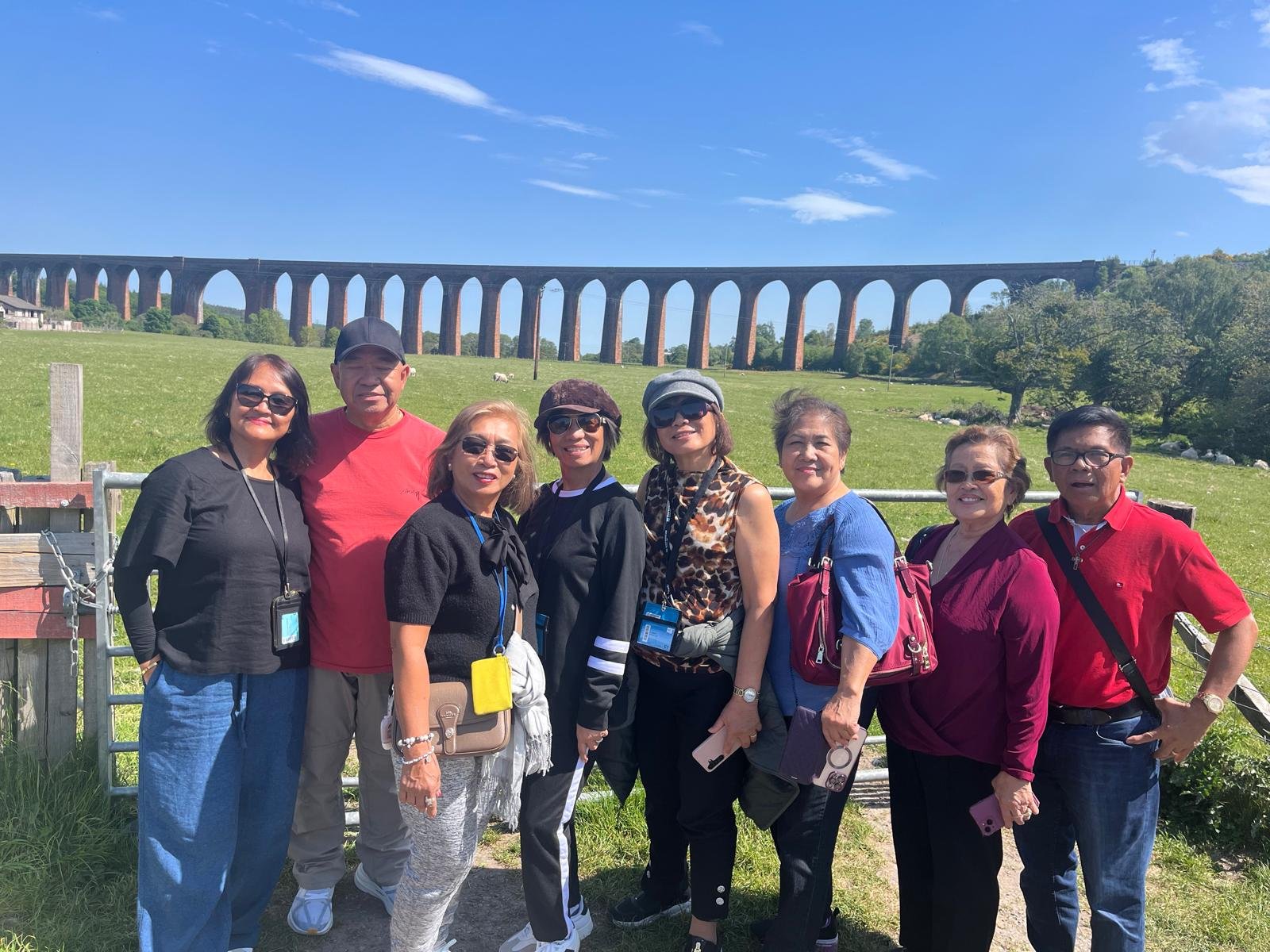 A group of nine diverse adults standing outdoors in front of a green field with trees and a large brick arched viaduct in the background, on a sunny day.