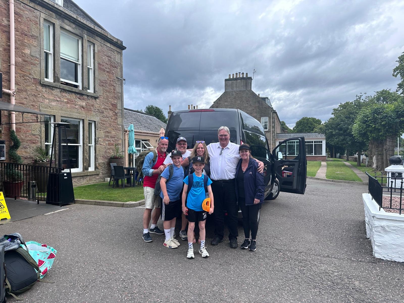 A group of eight people, including two children, standing in front of a black van outside a stone building. It is cloudy with overcast skies.