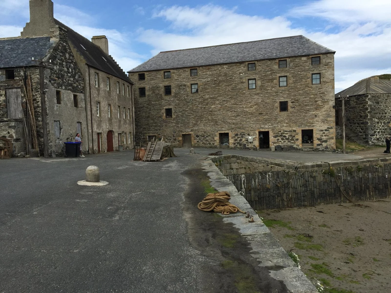 Old stone buildings with small windows, a cobblestone wall, and a sandy beach area with rope, in a village setting.