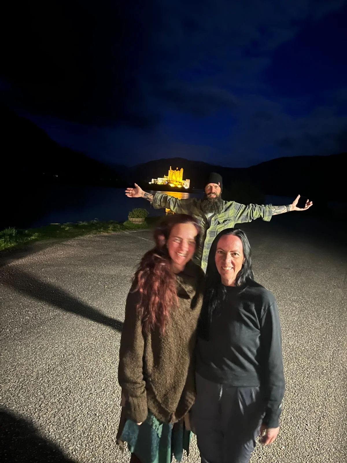 Three people standing on a gravel path at night, with a castle lit up in the background across a body of water. The person in the back has arms outstretched, while the two people in the front are smiling.