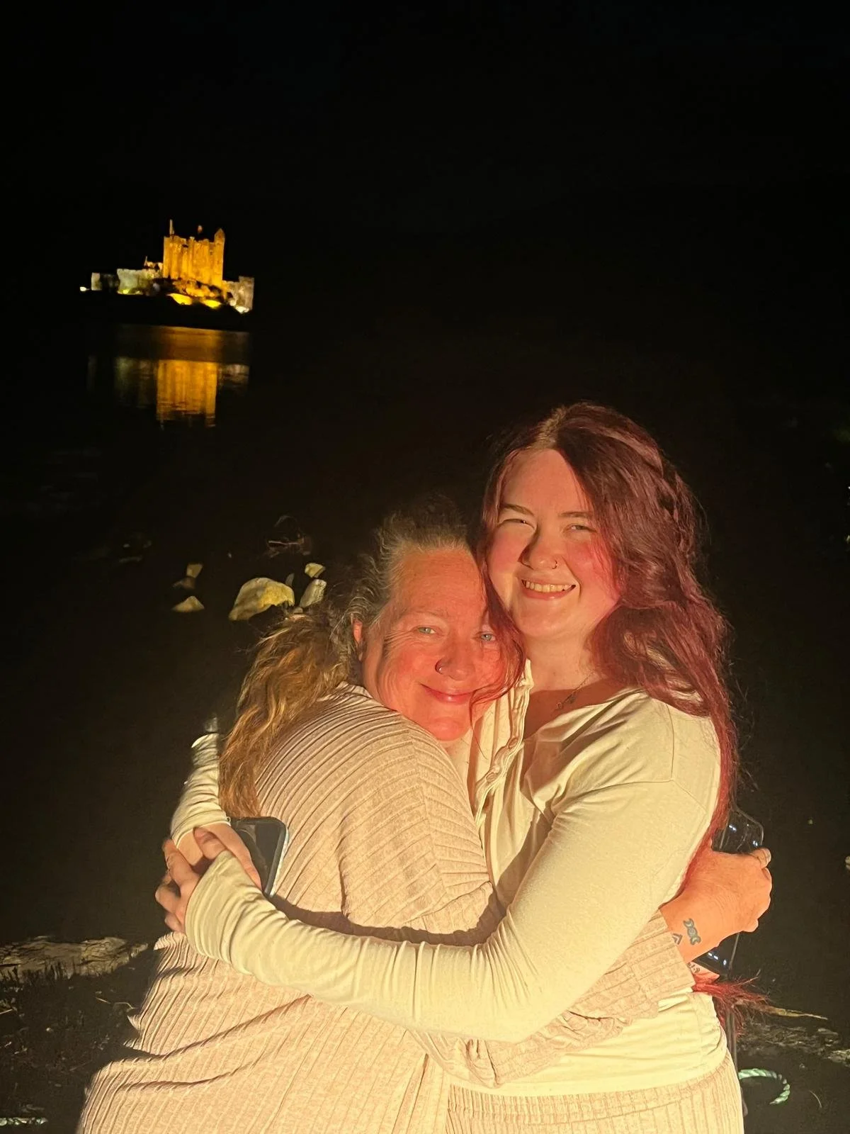 Two women hugging at night near a body of water with a castle illuminated in the background.