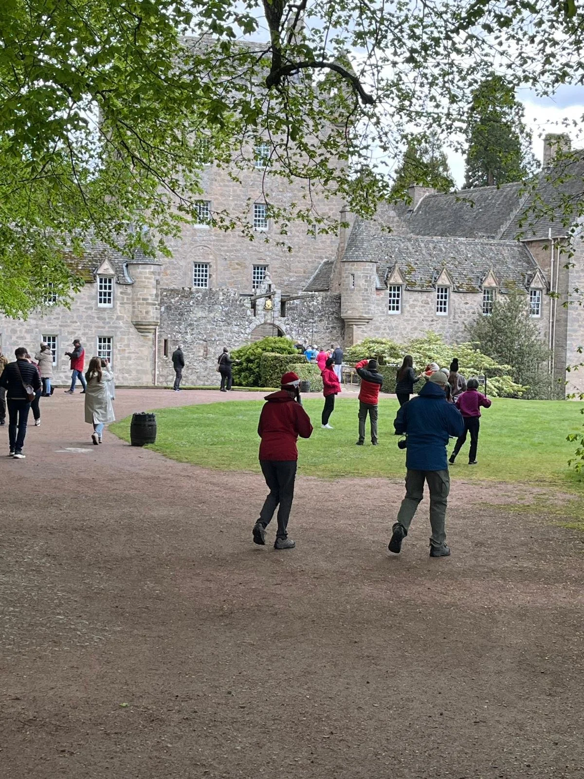 Tourists in front of a historic stone castle with multiple small windows and a tower, some taking photos and others enjoying the scenery, surrounded by green trees and grass, overcast sky.