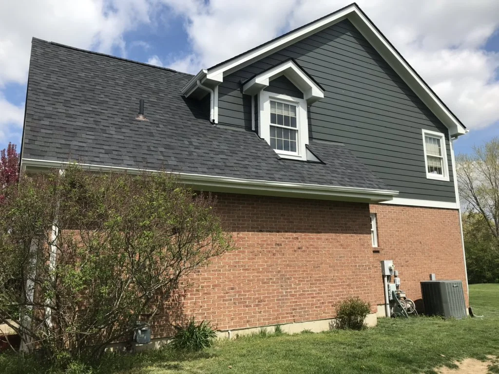 Side view of a two-story house with a brick lower level and dark gray siding upper level, with white trim around windows, and a gabled roof with asphalt shingles, surrounded by grass and trees.