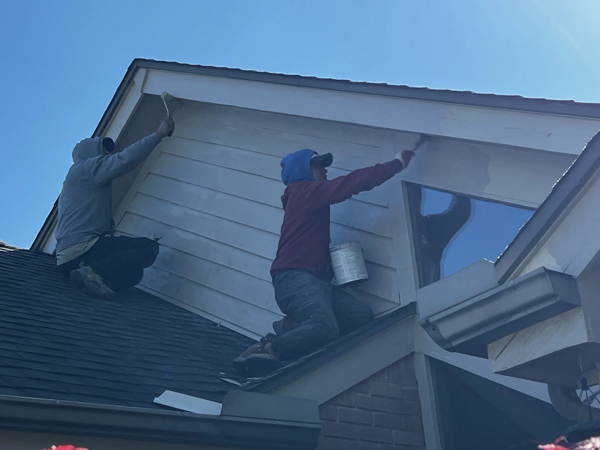Two workers on a ladder painting the exterior of a house with white paint under a clear blue sky.