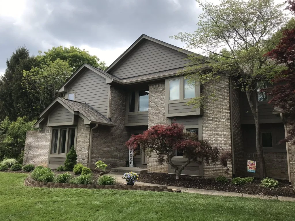 Large suburban house with brick and siding exterior, surrounded by a well-maintained garden with shrubs, trees, and flowers, under a cloudy sky.