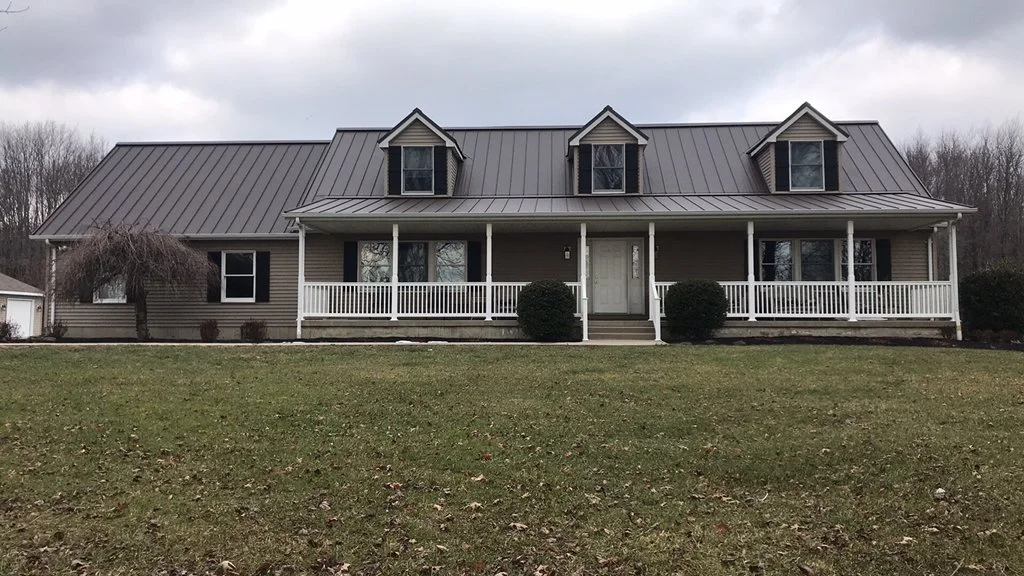 A large two-story house with beige siding, a gray metal roof, and three dormer windows. It has a front porch with white railings and steps leading to the front door. There are two bushes flanking the stairs, a leafless small tree on the left, and a l