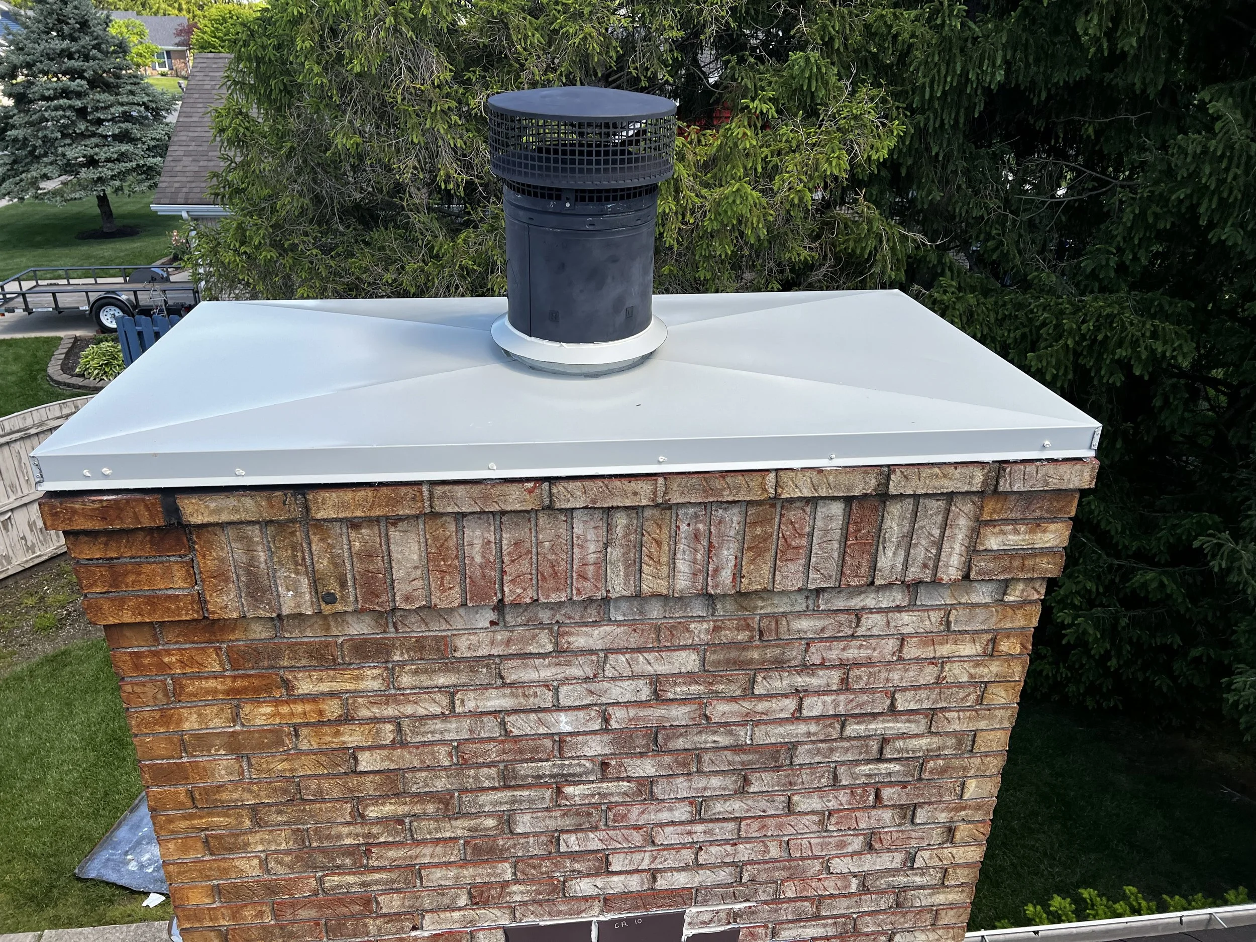 A chimney on a house roof with a black metal cap and a gray metal shield, surrounded by green trees and a lawn.
