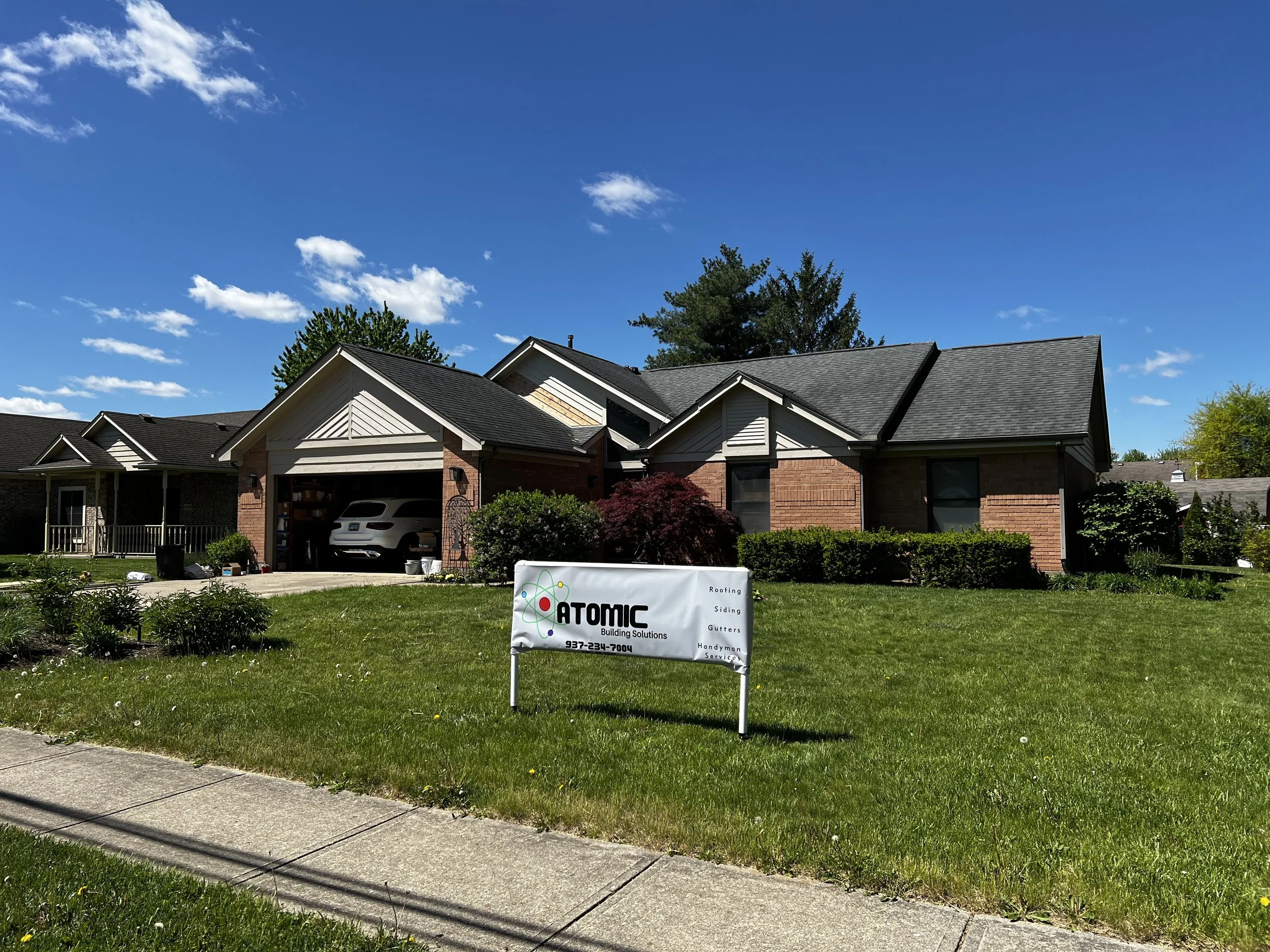 A residential house with a brick and siding exterior, multiple gable roofs, and a driveway with a garage. There is a green lawn with a small tree and bushes in front. A sign in the yard advertises Atomic Building Solutions, including roofing, siding,