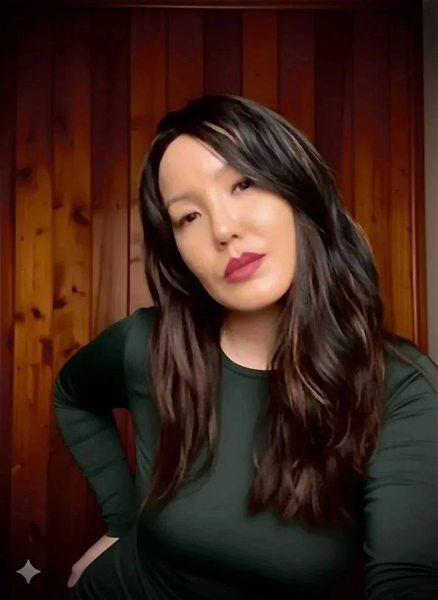 Mulher com cabelo escuro, longo e ondulado, usando blusa verde escura, posando na frente de um painel de madeira