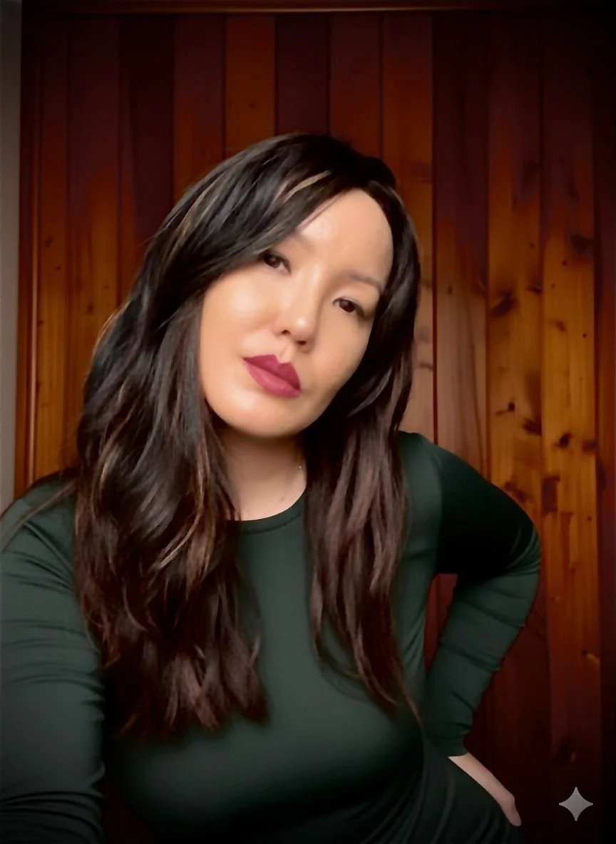 Mulher de pele clara, com cabelo castanho escuro e ondulado, usando batom vinho escuro, vestindo uma blusa verde, posando com uma parede de madeira ao fundo.