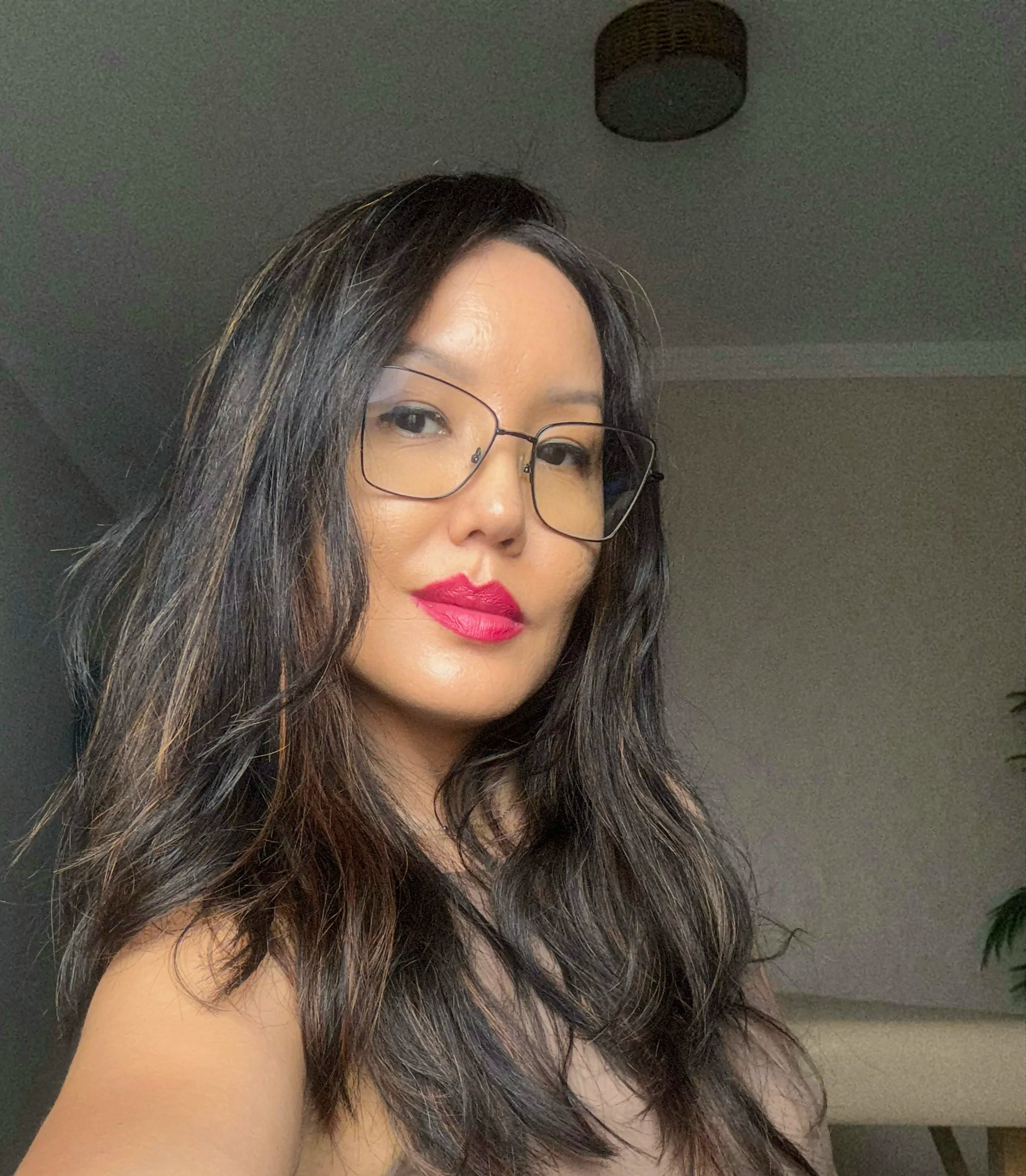 Mulher com cabelo castanho escuro e ondulado, óculos de armação fina e maquiagem com batom rosa, posando para a câmera em ambiente interno.