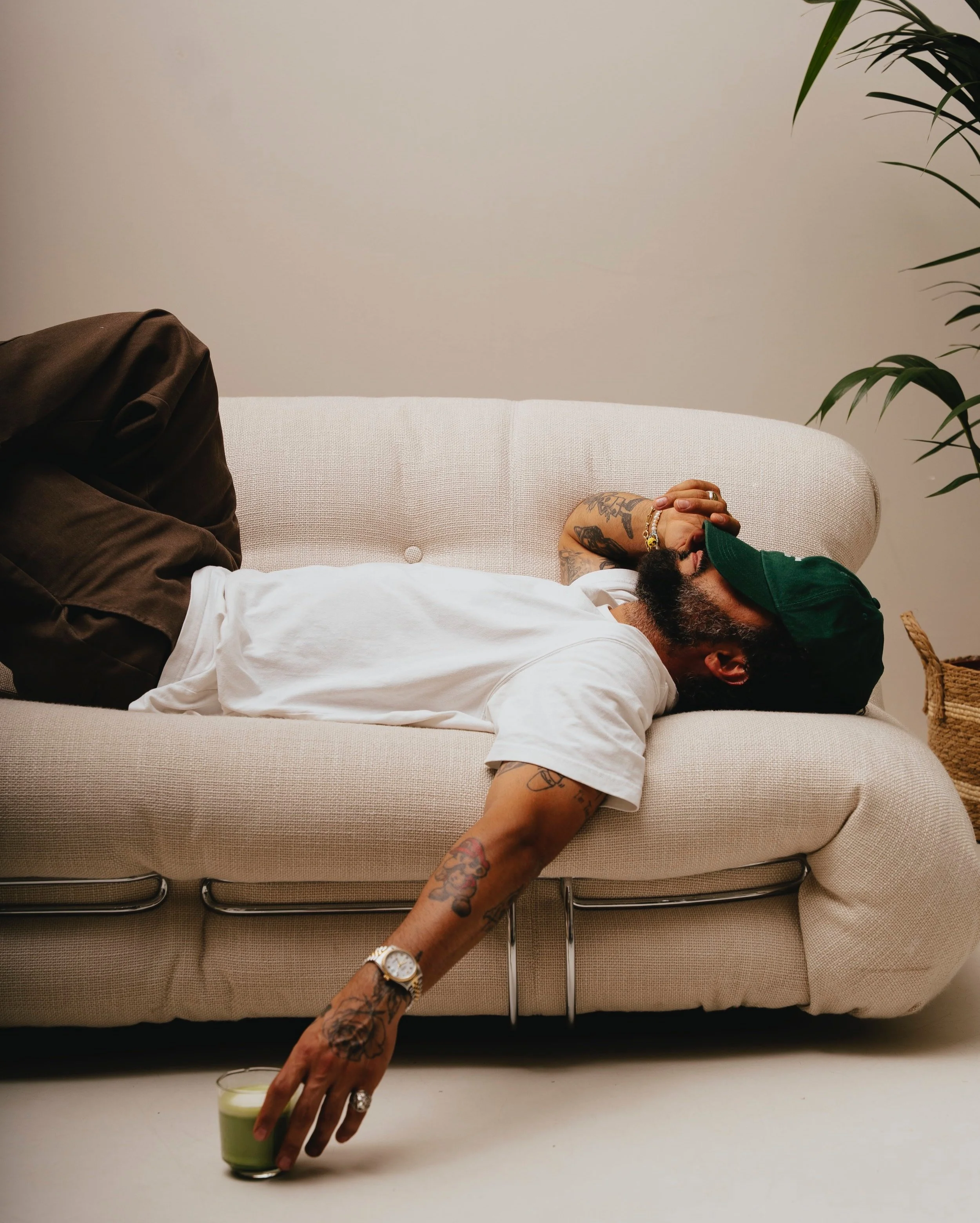 Ein Mann liegt auf einem weißen Sofa, trägt ein weißes T-Shirt, eine grüne Cap und hat Tattoos auf Arm und Hand. Er hält ein Glas grünen Saft. Der Mann sieht müde aus und hat die Hand vor das Gesicht. Im Hintergrund ist eine grüne Pflanze zu sehen.