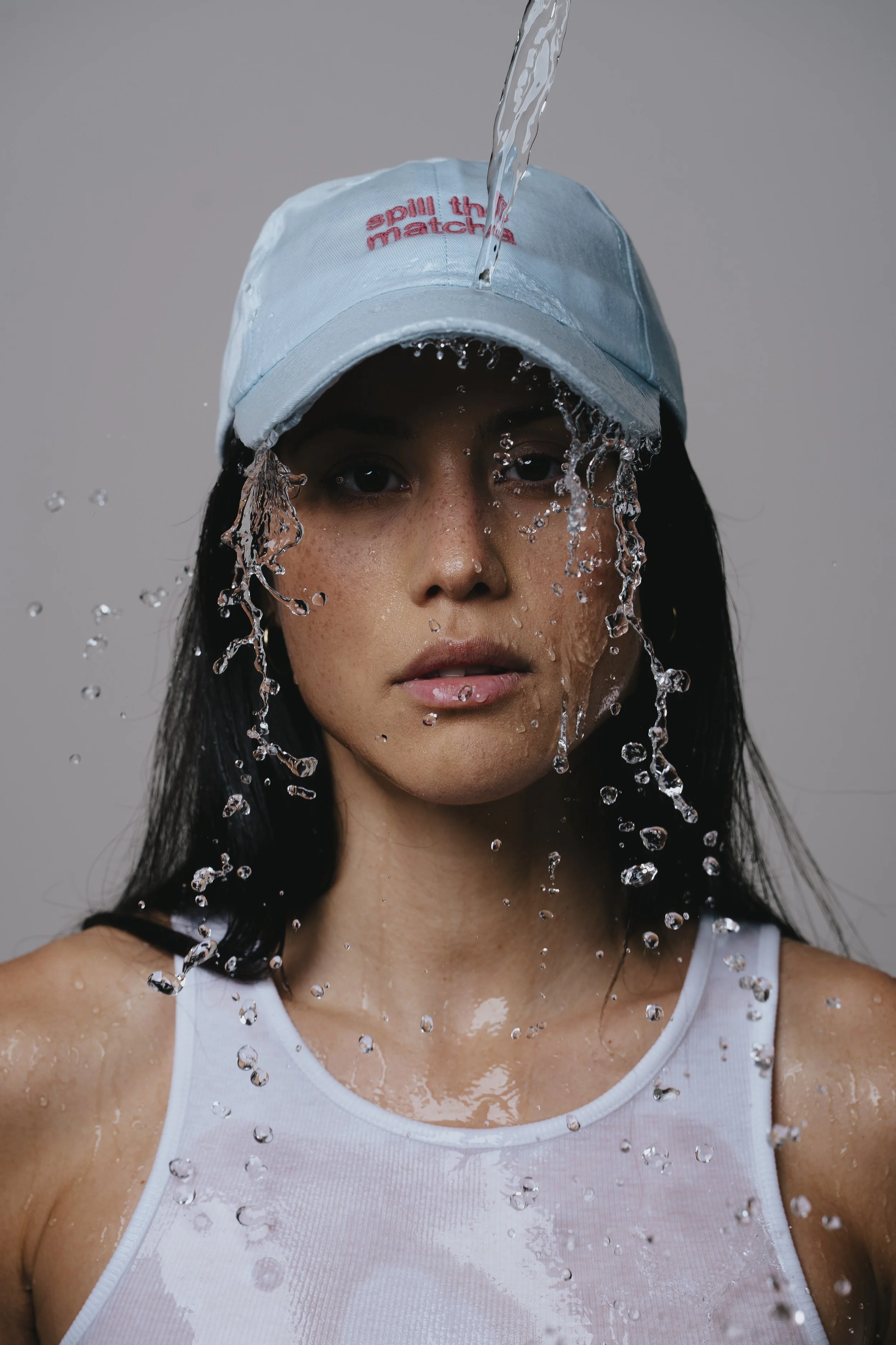 Junge Frau mit nassem weißen Tanktop und blauer Baseballkappe leckt Wasser aus dem Hut, Wasser tropft von ihrem Gesicht.