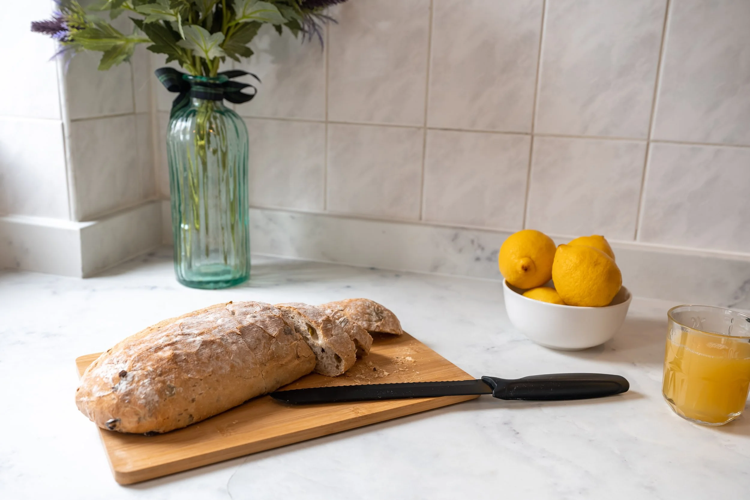 Fresh loaf of bread on a wooden cutting board with a bread knife, a bowl of lemons, a glass of orange juice, and a vase with flowers on a marble kitchen countertop.