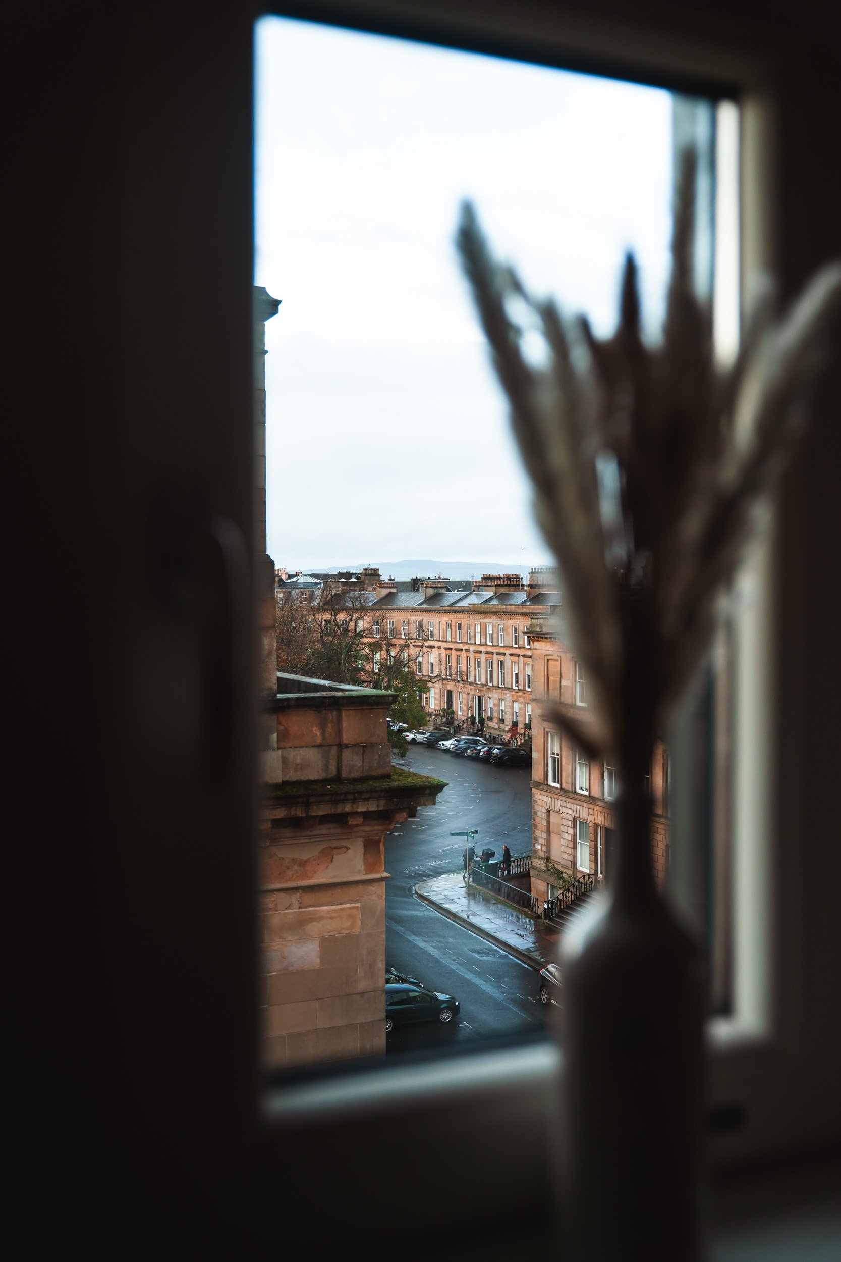View of a city street through a window with a blurry vase of dried flowers in the foreground.