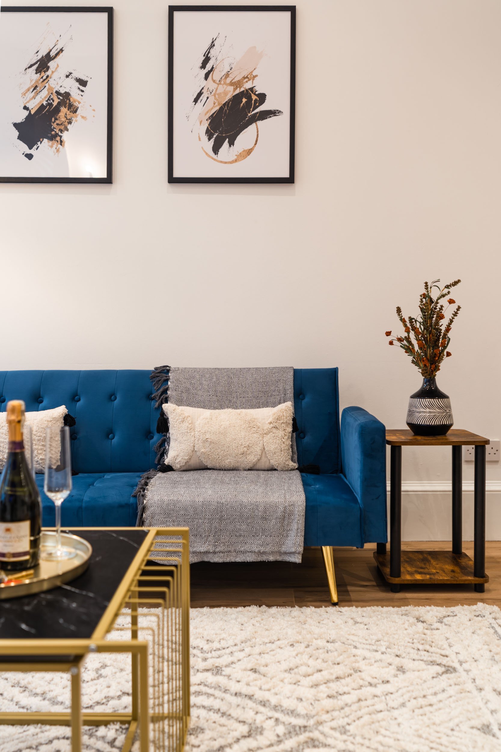 A living room with a blue velvet couch, beige and white pillows, a gray throw blanket, framed abstract art on the wall, a floral arrangement in a black and white vase on a small wooden table, and a gold-framed coffee table with a bottle of champagne 