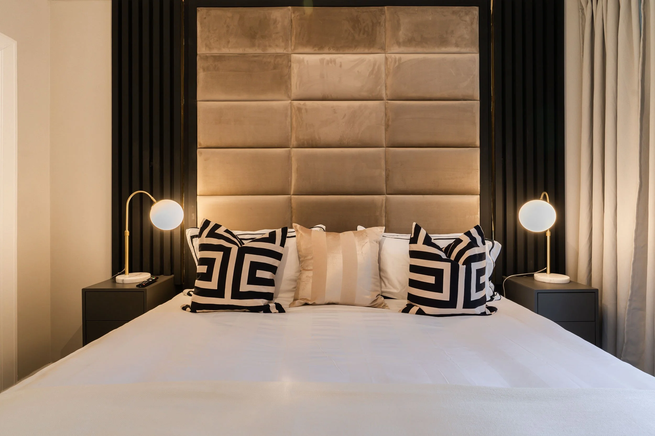 A modern bedroom featuring a large bed with black and white geometric patterned pillows, a beige striped pillow, and two bedside tables with white round lamps on either side.