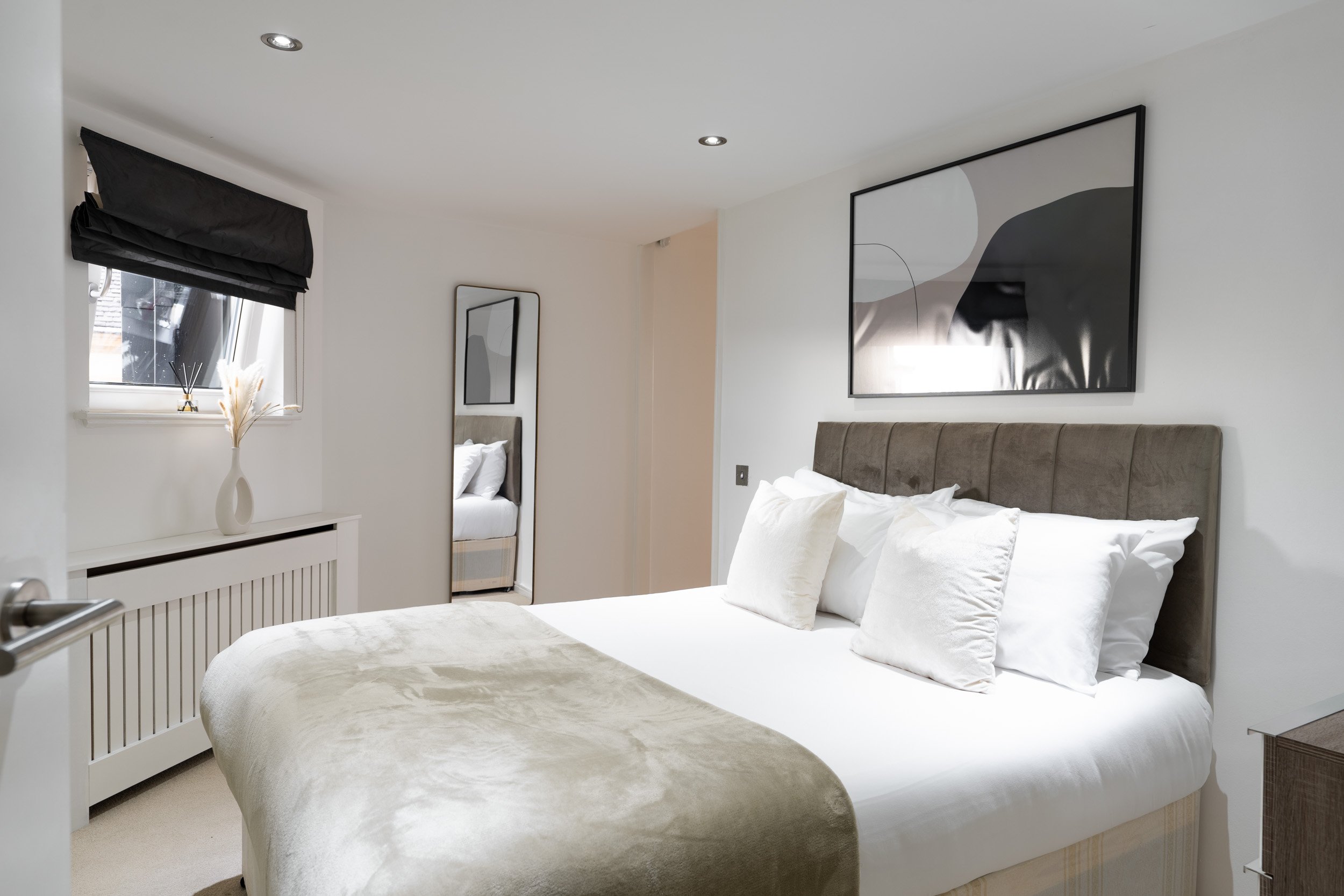 A neatly made bed with white pillows and beige blanket in a modern bedroom with abstract wall art and a full-length mirror.