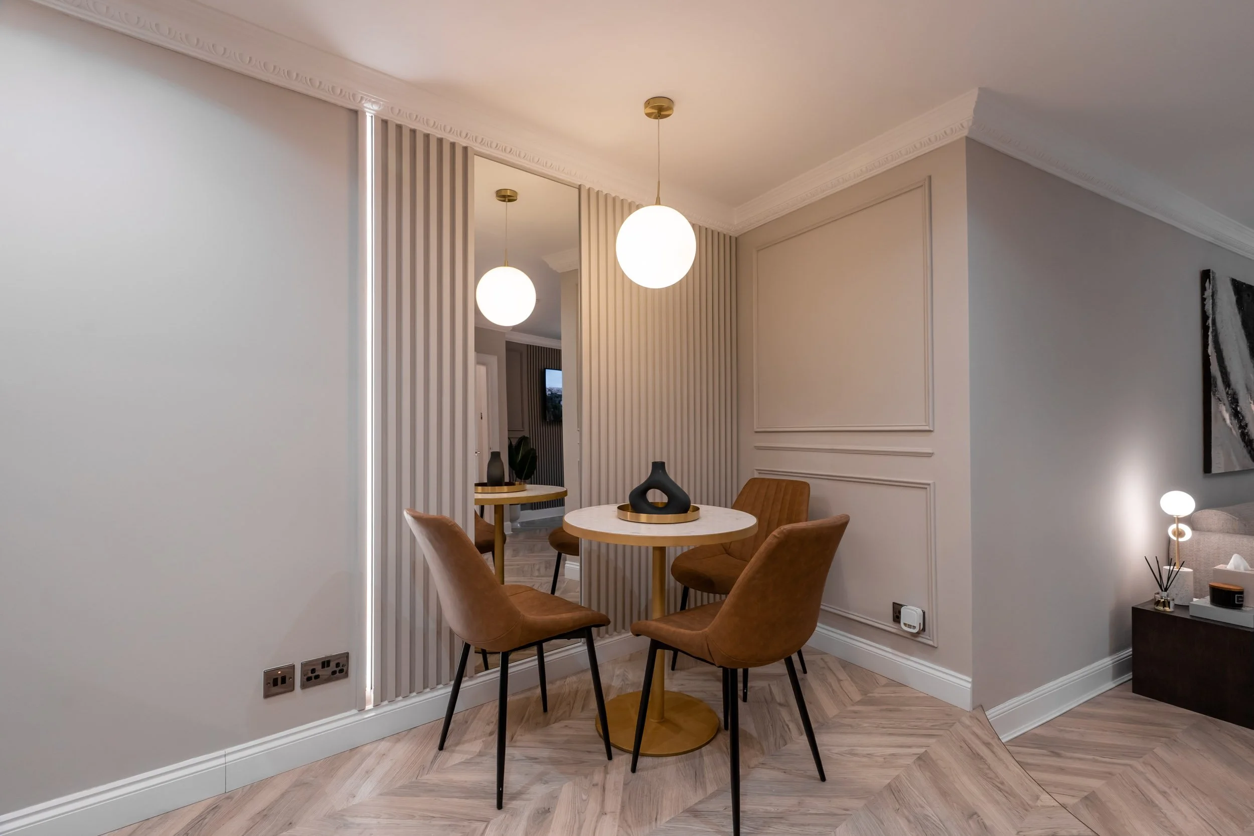 A modern dining area with a round table, four brown upholstered chairs, and decorative black vases on the table. Two pendant lights hang from the ceiling, and there's a mirror on the wall reflecting part of the room. The floor has a herringbone patte