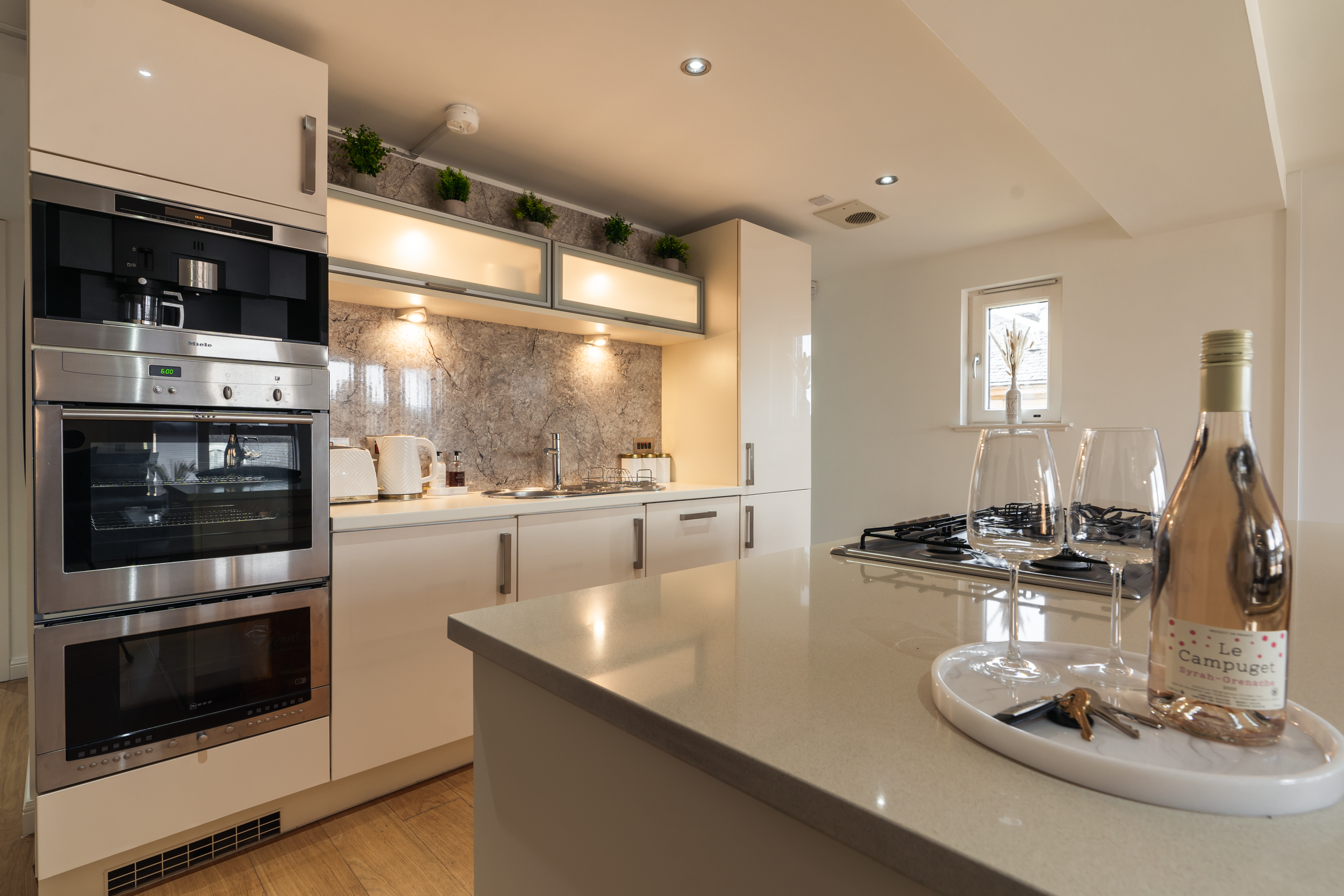 Modern kitchen with cream-colored cabinets, a built-in oven, and a stove on the countertop. A small window with a vase of dried plants, and a white tray on the island with a bottle of rosé and two wine glasses.