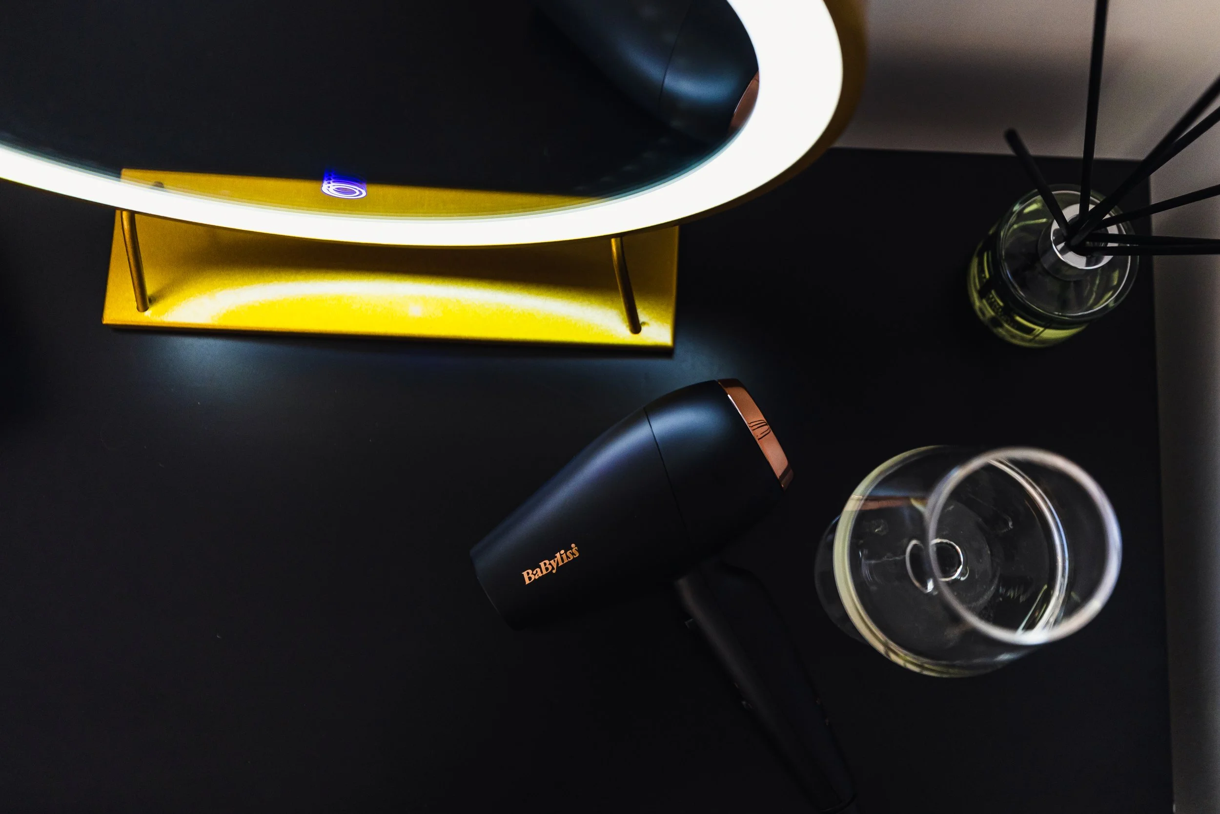 Overhead view of a black and copper BaByliss hair dryer, a glass of water, a glass diffuser, and a modern lamp with a white and black shade on a dark surface.
