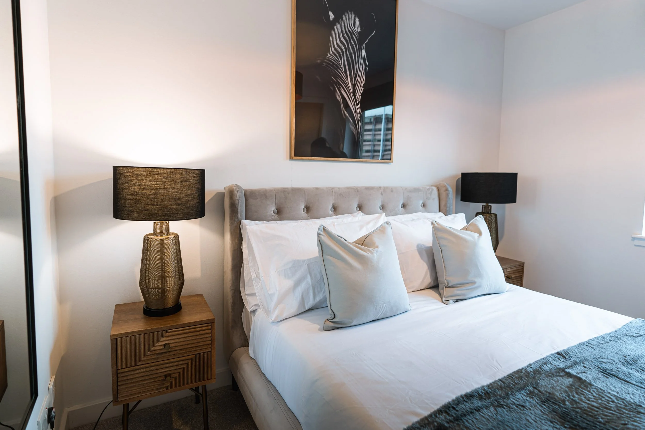 A bedroom with a bed, beige upholstered headboard, white bedding, two beige pillows, and two black lamps on wooden nightstands. There is a framed zebra artwork on the wall above the bed.