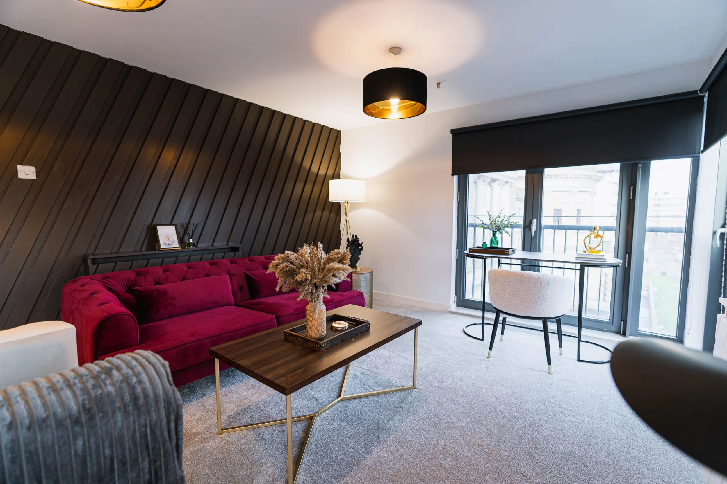 Modern living room with a deep red velvet sofa, a wooden coffee table, and a large window with black blinds. Decor includes a vase with dried flowers on the coffee table, a framed picture, and a small decorative statue on a side table. There is a flo