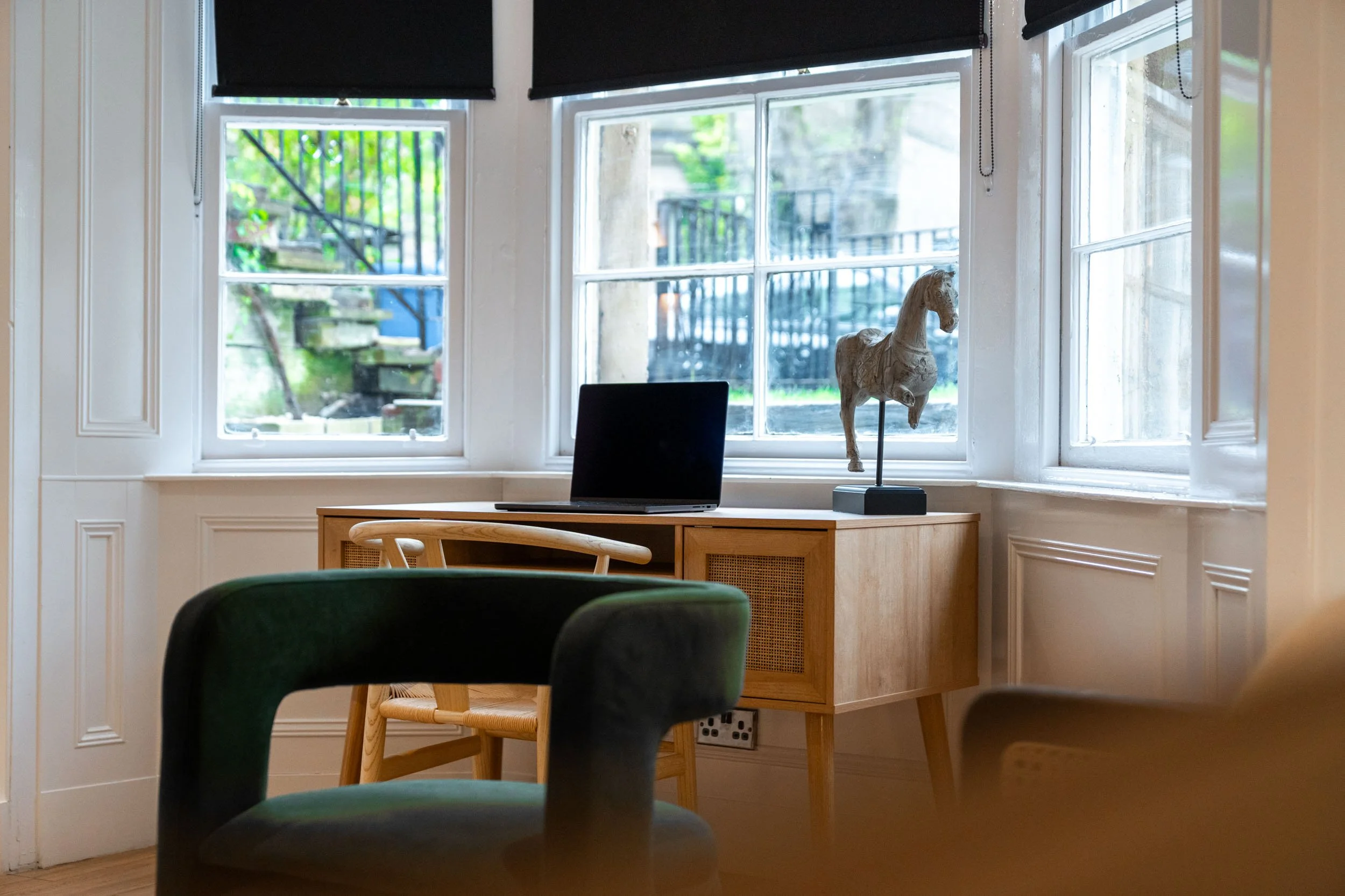 A cozy room with a wooden desk and a laptop on it, positioned in front of three large windows with black blinds. A decorative horse sculpture is on the desk, with one window showing an outdoor staircase and greenery.