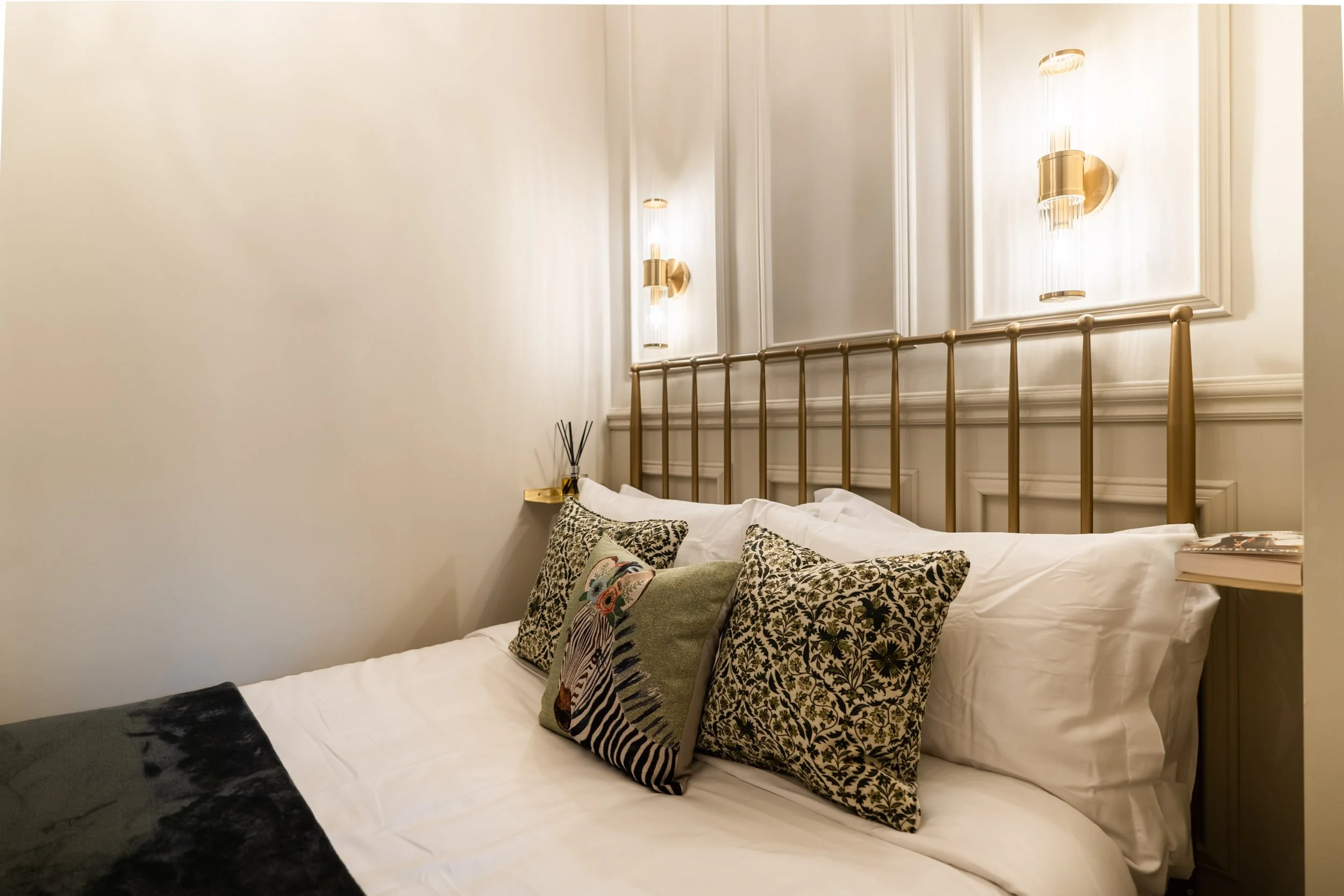 A neatly made bed with white sheets, decorative pillows with floral and animal print, and a black throw blanket at the foot, against a white wall with gold wall sconces.