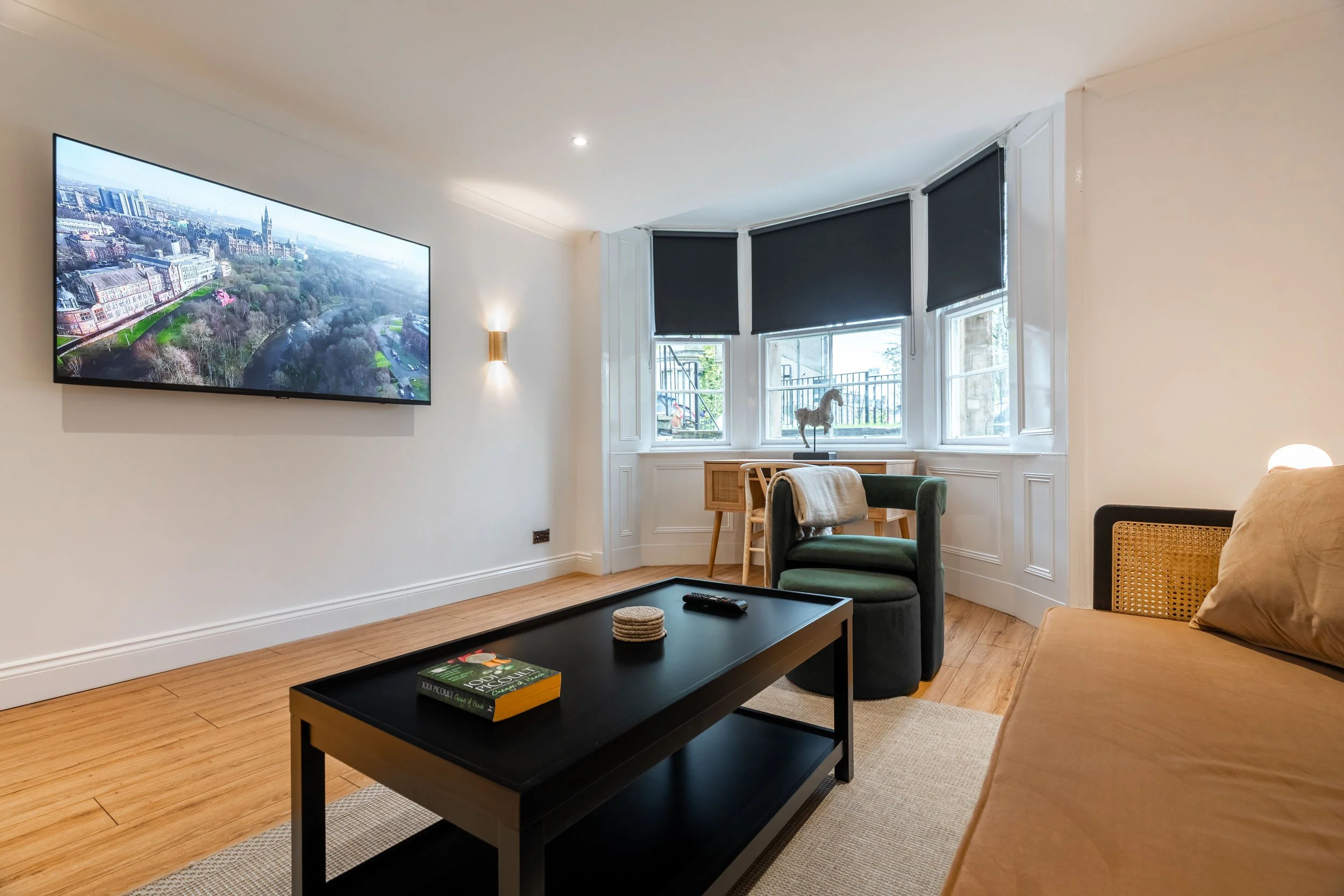 Living room with wall-mounted TV, green armchair, wooden desk with horse figurine, beige sofa, black coffee table with book and remote, large bay window with black blinds.