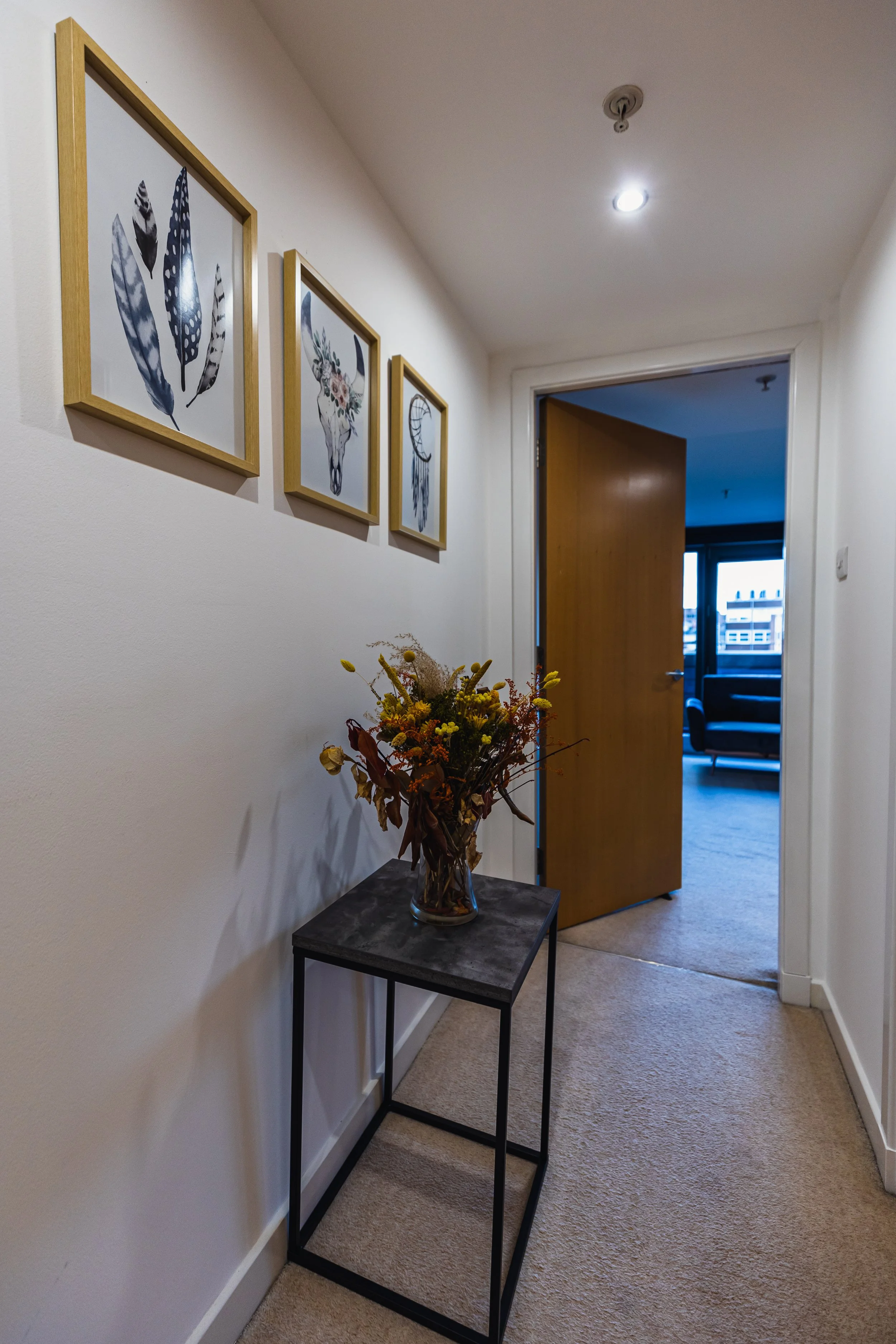 A hallway with a small table holding a dried flower arrangement, three framed pictures of feathers and animals on the wall, an open door revealing a living room with large windows, a chair, and a sofa.