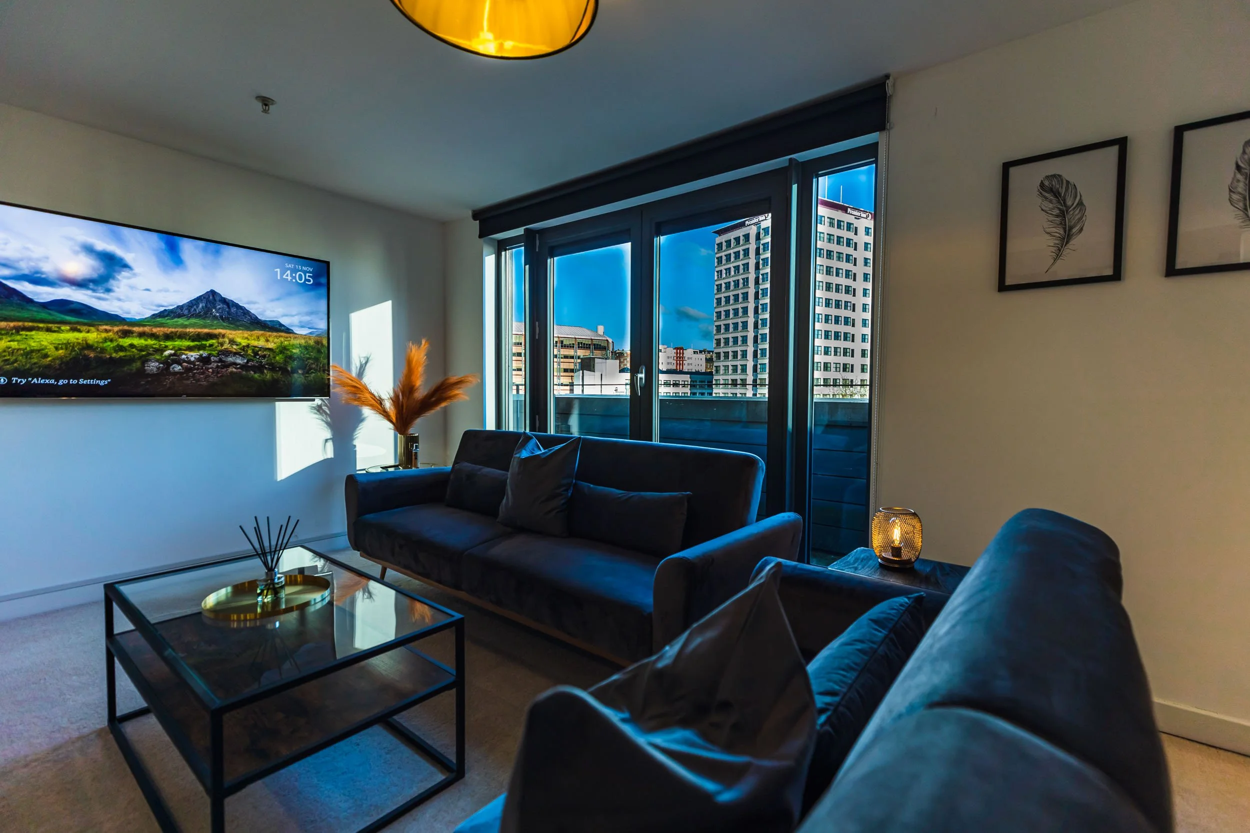 Modern living room with dark sofa, glass coffee table, TV on wall, large windows showing city skyline, framed feather art, and decorative plants