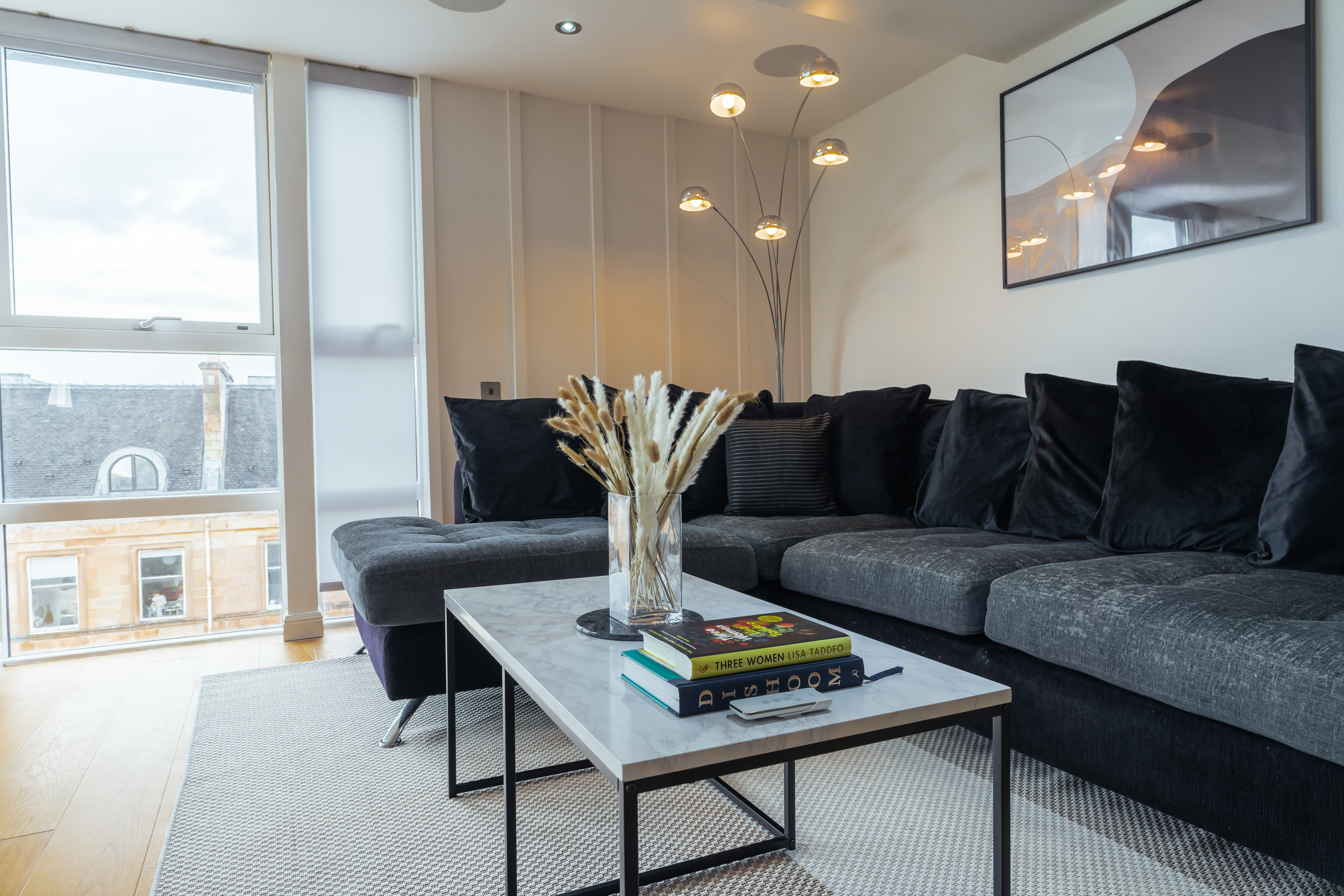 Modern living room with a gray sectional sofa, black pillows, a white coffee table with books and a vase of dried plants, large windows with blinds, a multi-head floor lamp, and a framed artwork on the wall.