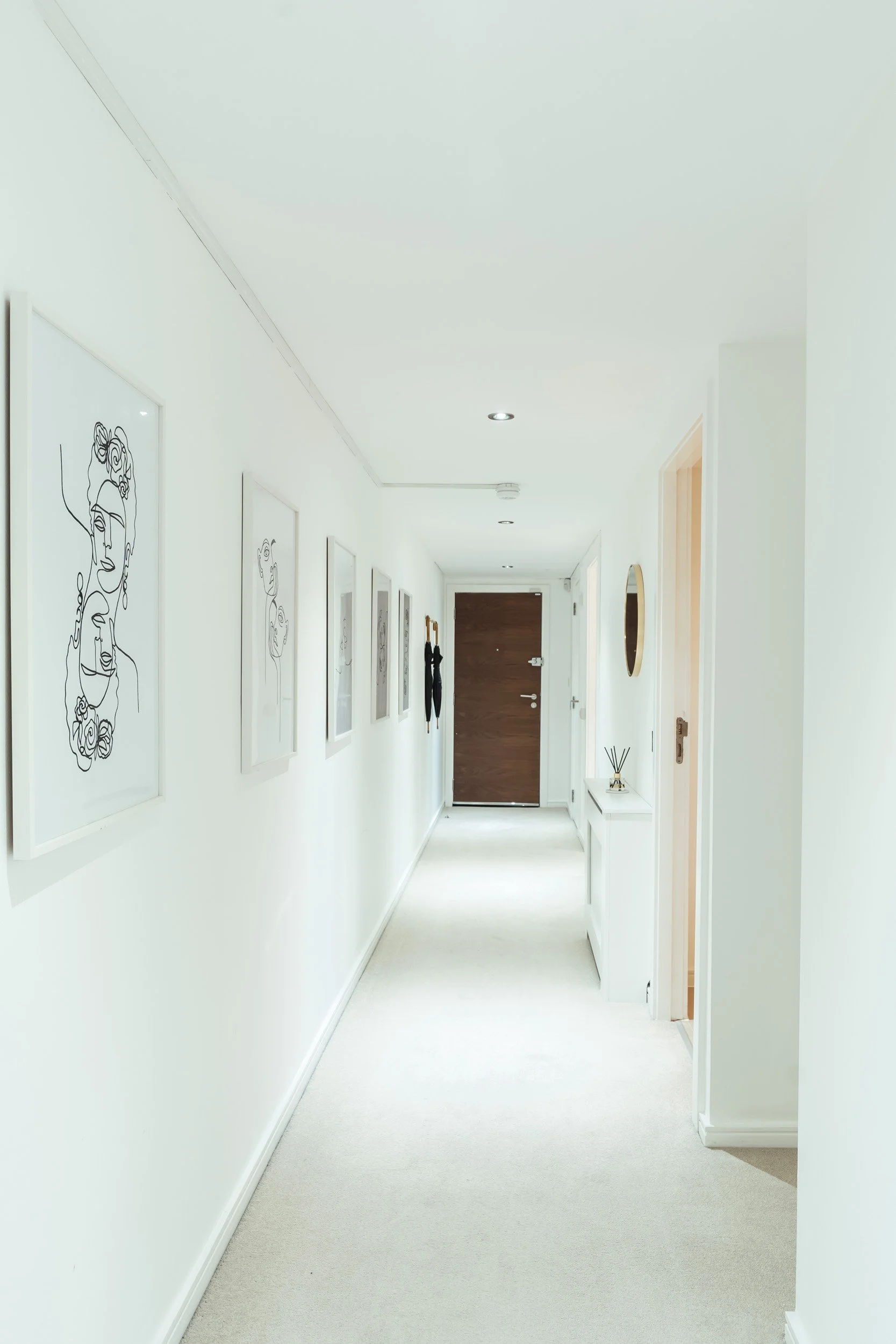 White hallway with framed art on the left wall, a small white table with a reed diffuser, and black umbrellas hanging near a wooden door at the end.