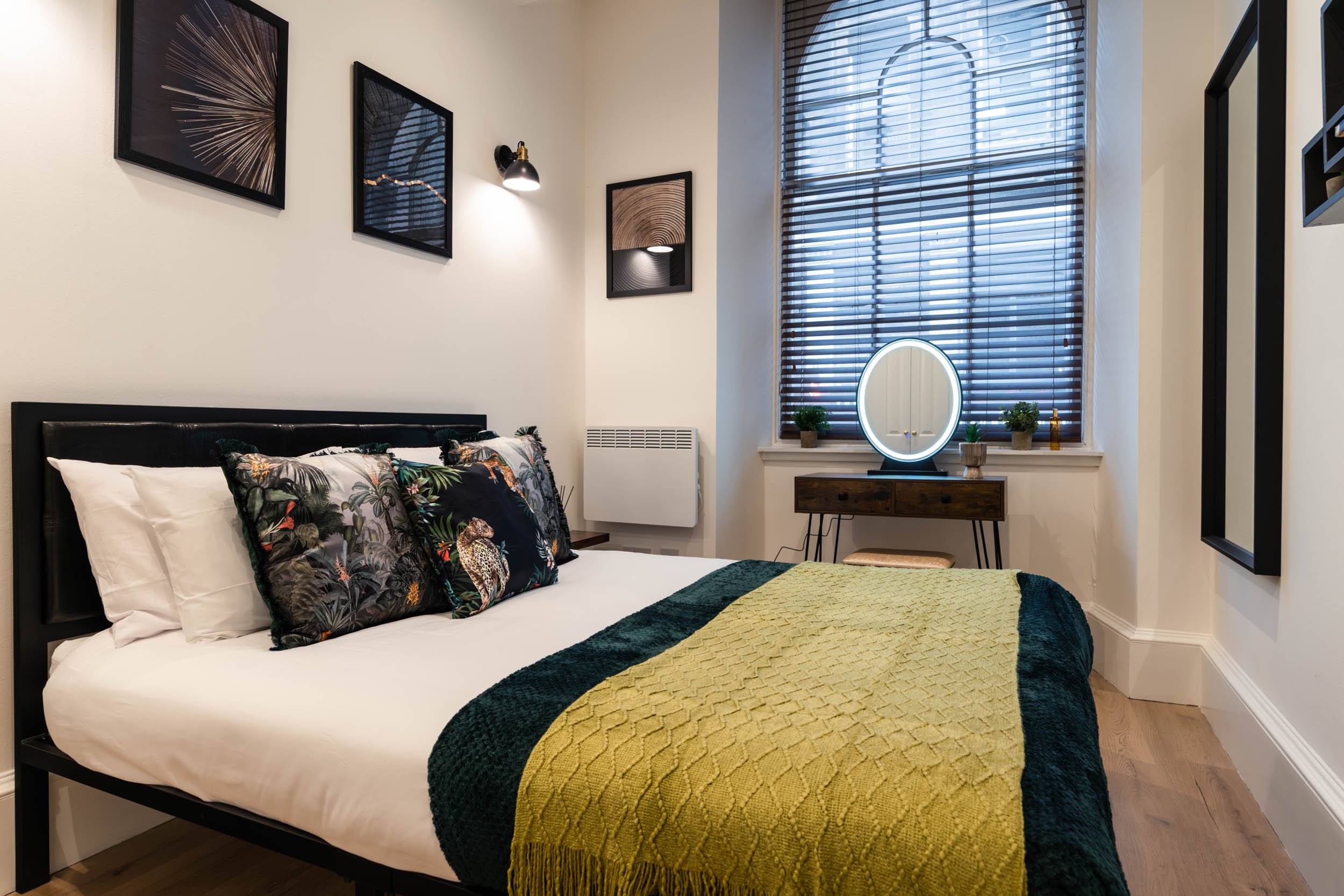 A bedroom with a black bed frame, decorated with tropical-themed pillows, a yellow and green blanket, and artwork on the wall. There's a window with wooden blinds, a small wooden vanity with a round mirror, and small potted plants on the windowsill.