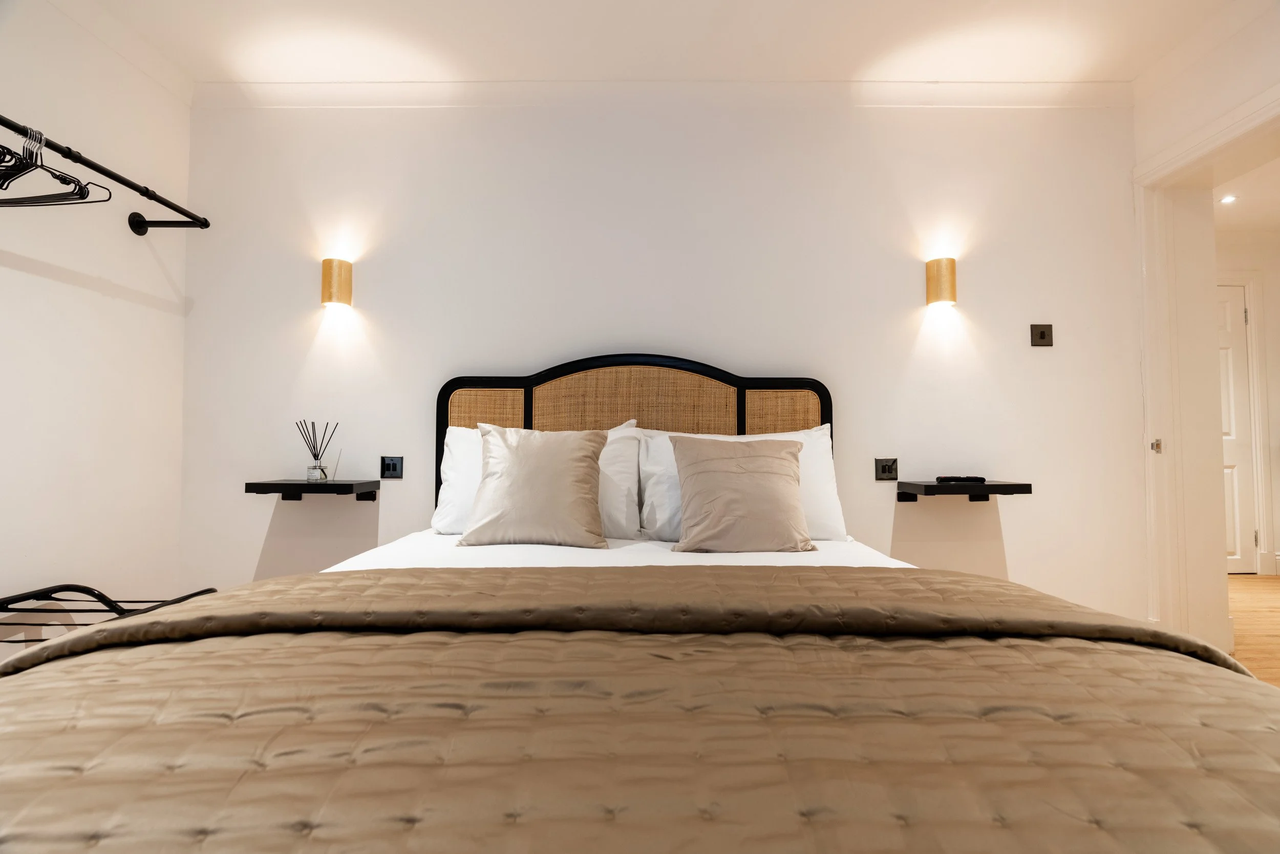 A neatly made bed with beige pillows, white sheets, and a beige quilt in a well-lit bedroom with modern light fixtures on the wall. There are small black floating bedside shelves on each side of the bed.