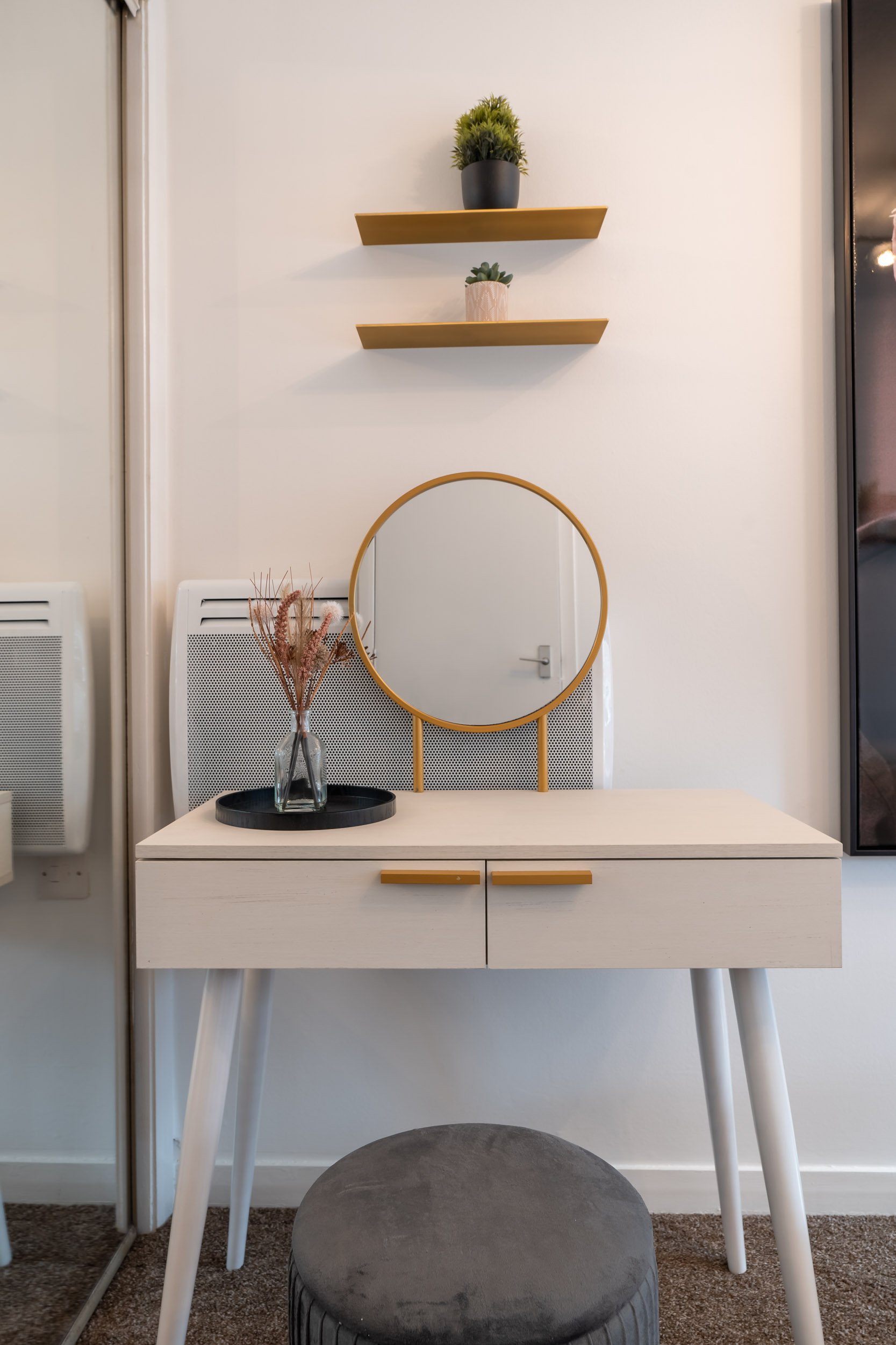 A white vanity table with a circular mirror, a black tray with dried flowers, and a decorative stool in front. Two floating shelves with plants are on the wall above.