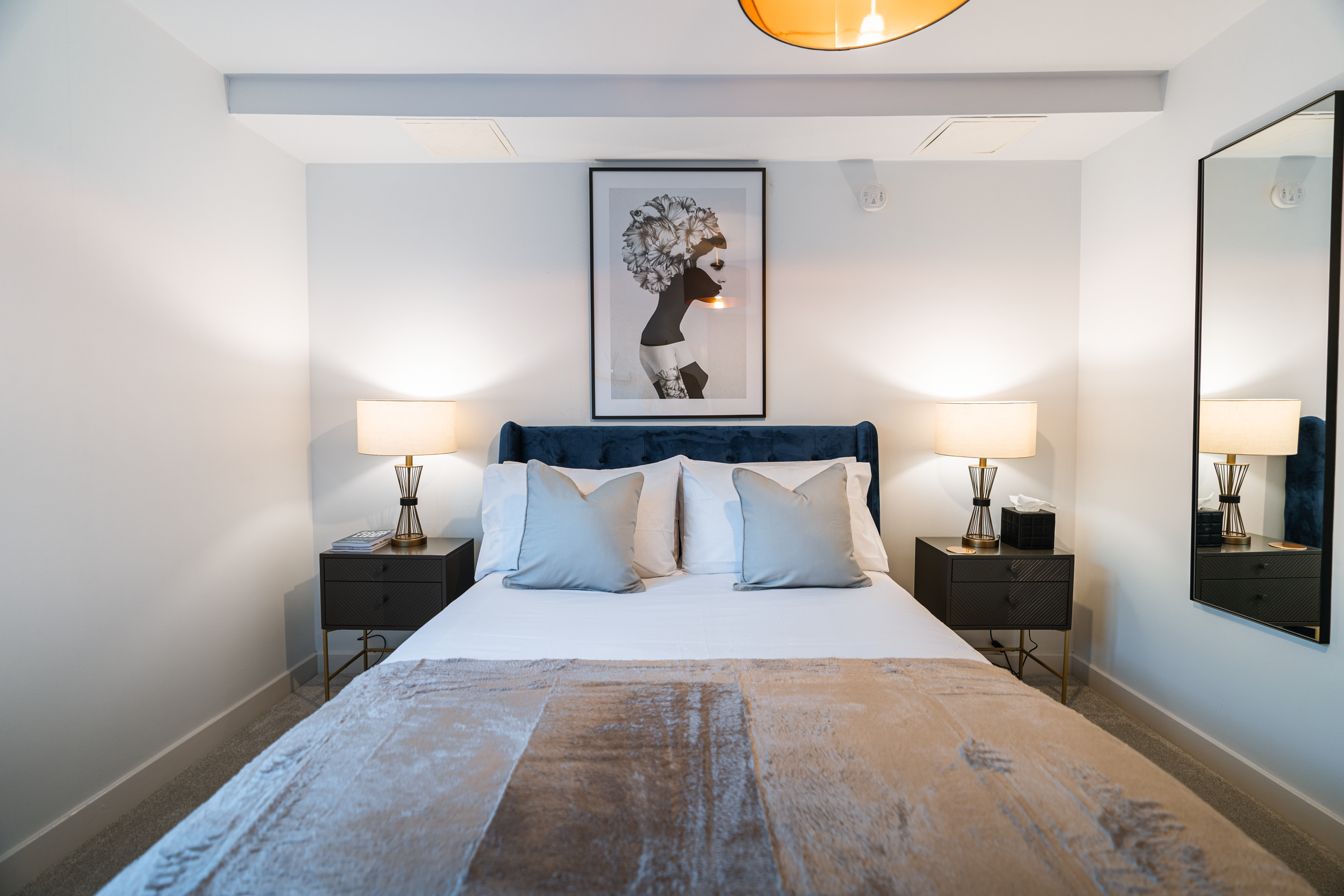 A modern bedroom with a white bed, four pillows, a blue velvet headboard, two black nightstands with lamps, a large mirror on the right wall, and a framed black and white art piece above the bed.