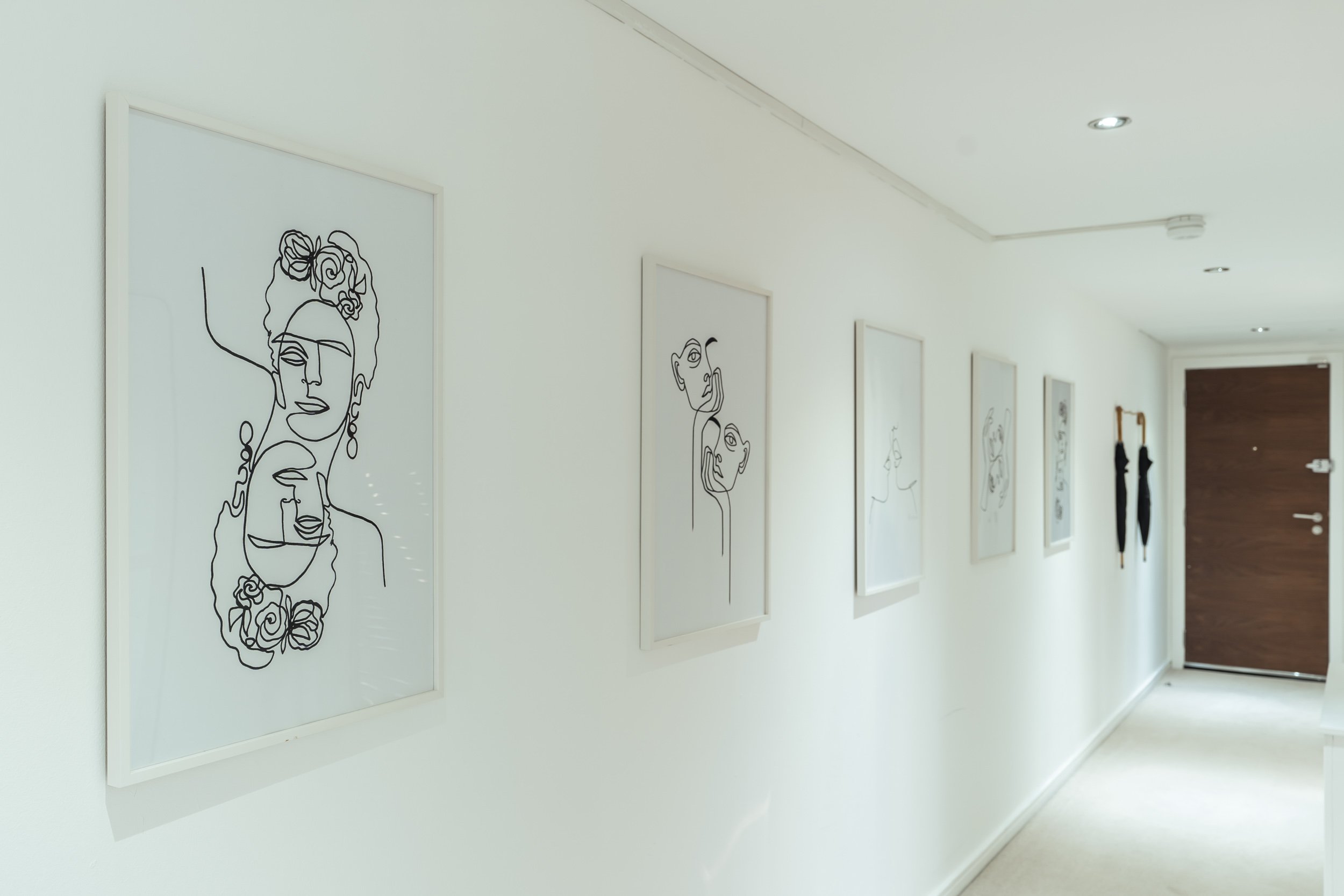 A white hallway with framed line art portraits of women and men on the left wall, with a wooden door and black umbrellas hanging on the wall near the door at the end of the hallway.