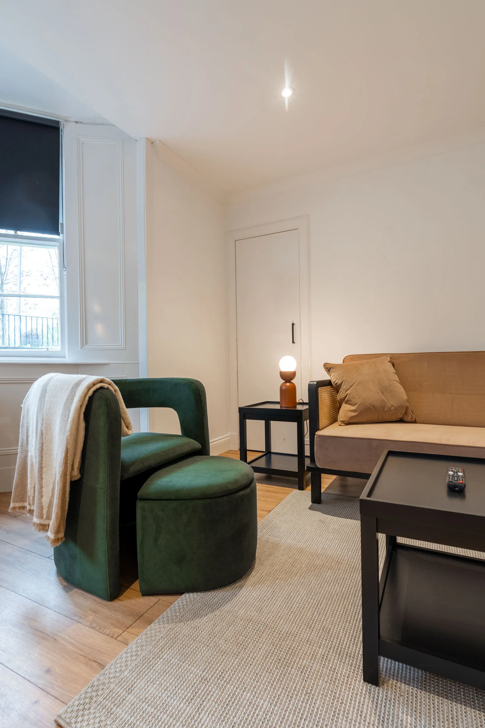 Living room with green armchair, beige couch, black side table with lamp, TV tray table with remote, window, hardwood floor, and neutral-colored rug.