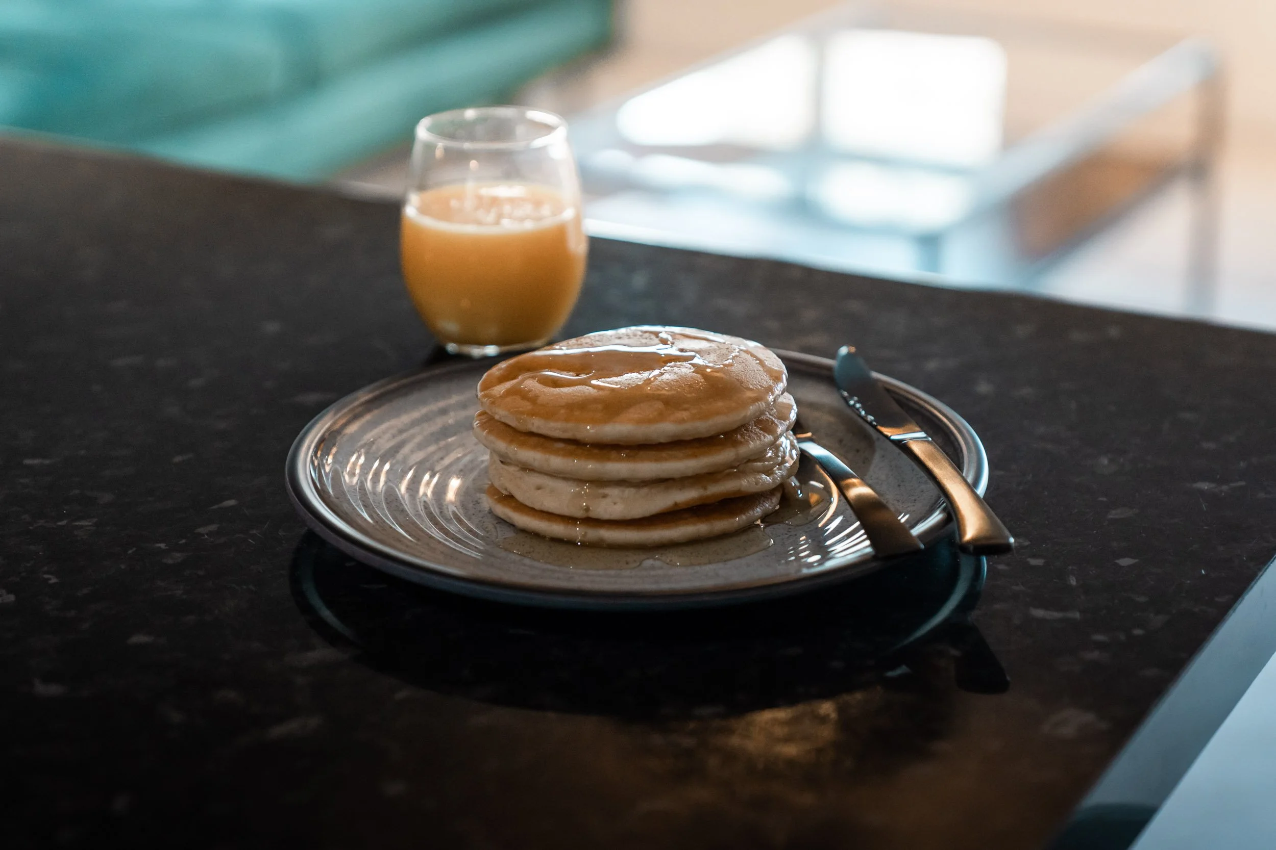 A plate of four pancakes drizzled with syrup, a butter knife and fork on a black countertop, with a glass of maple syrup in the background.