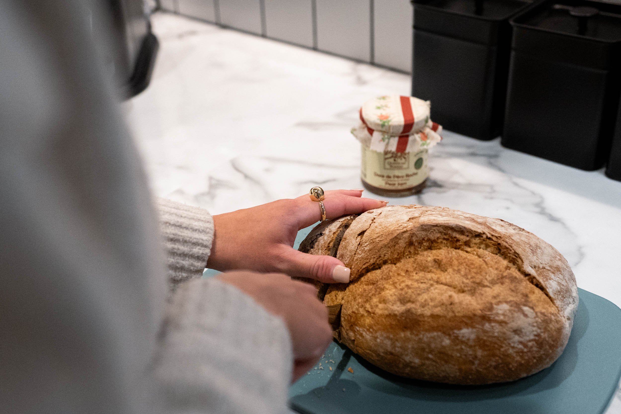 Person slicing a loaf of rustic bread on a blue cutting board in a kitchen with a jar of jam in the background.