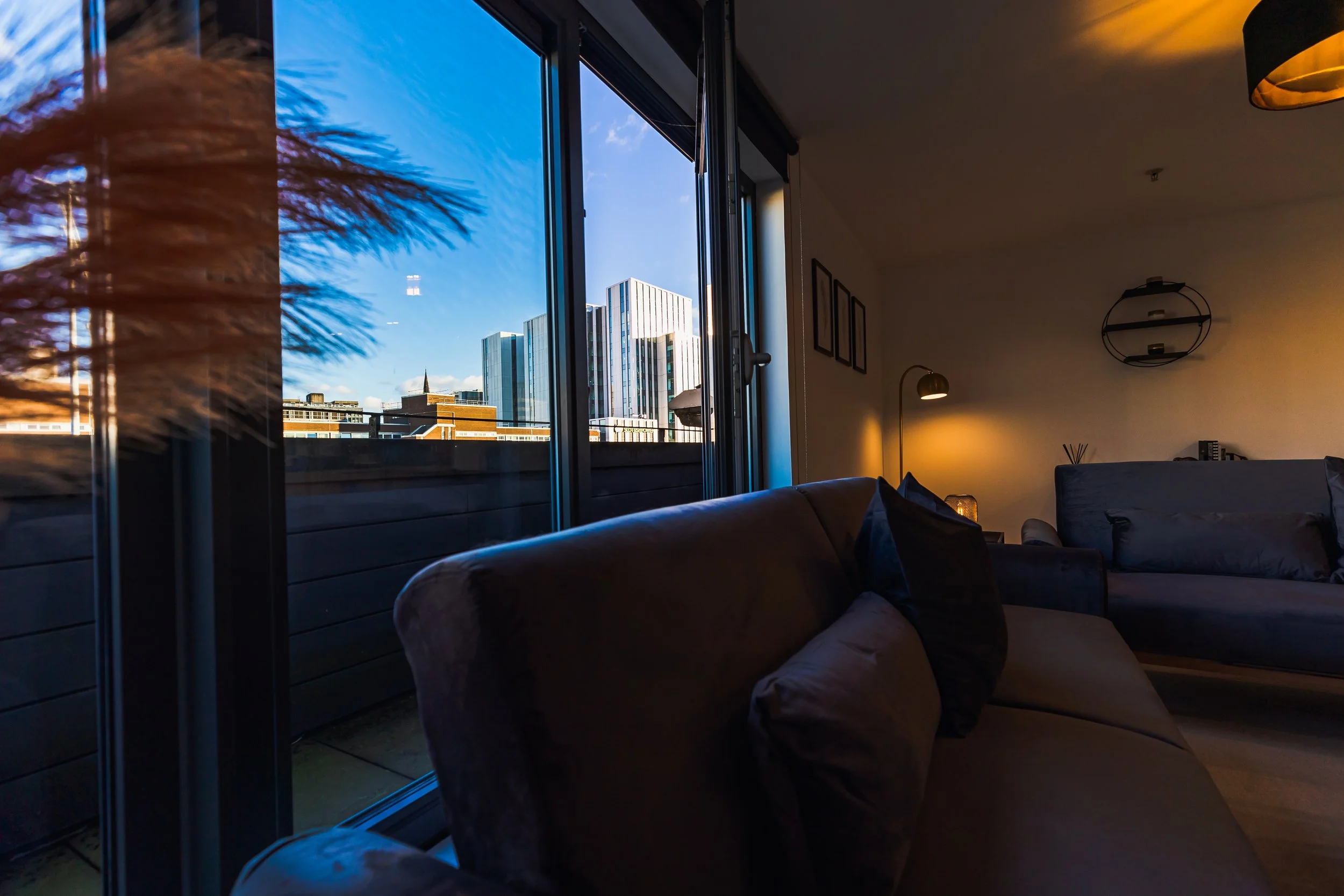 Interior of a modern living room with a dark sofa and pillows, illuminated by warm yellow lighting, with large glass sliding doors showing a city skyline with tall buildings against a blue sky.