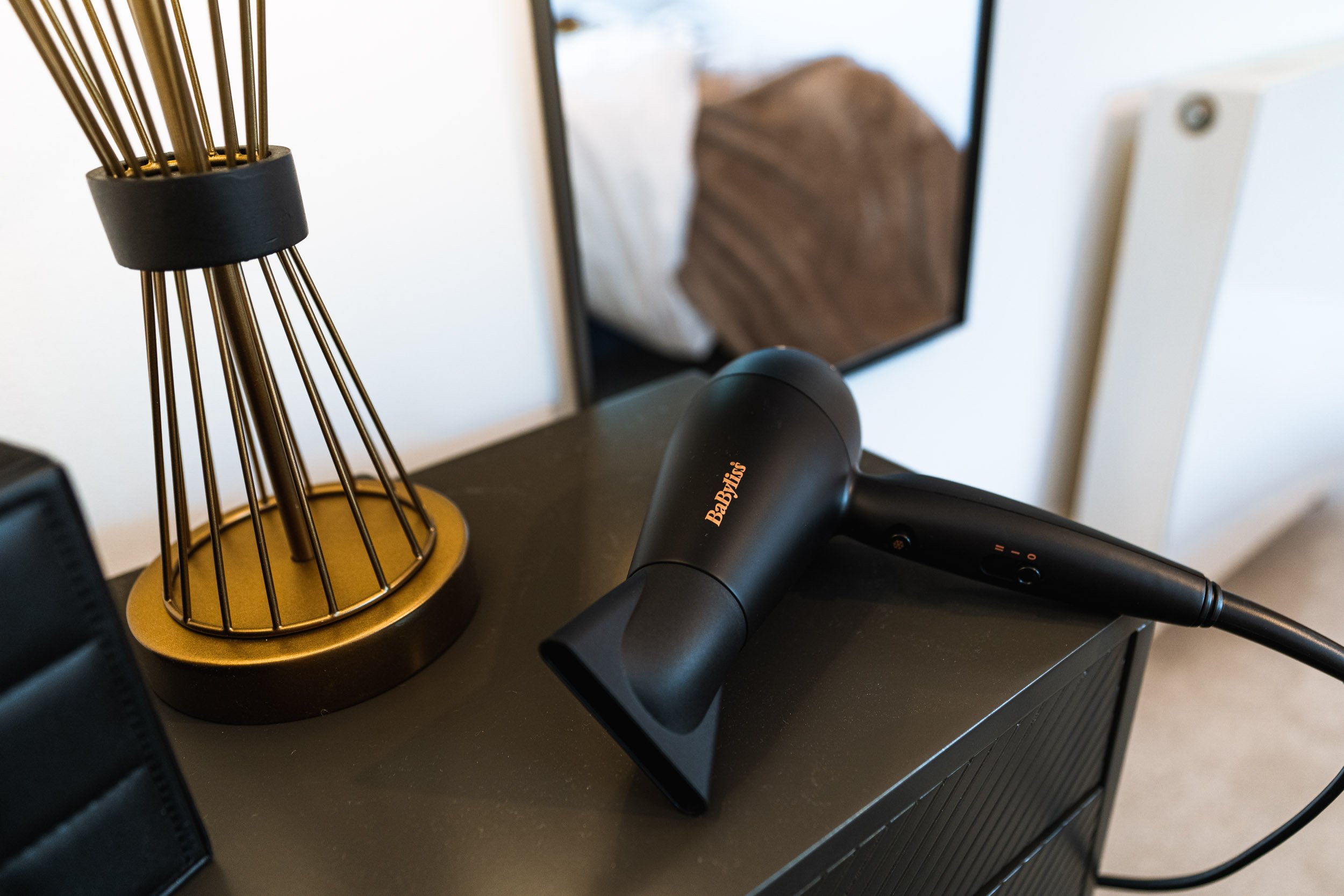 Black hair dryer with the brand name 'BaByliss' placed on a dark-colored surface next to a decorative gold and black wire lamp and a mirror, with a white wall and radiator in the background.