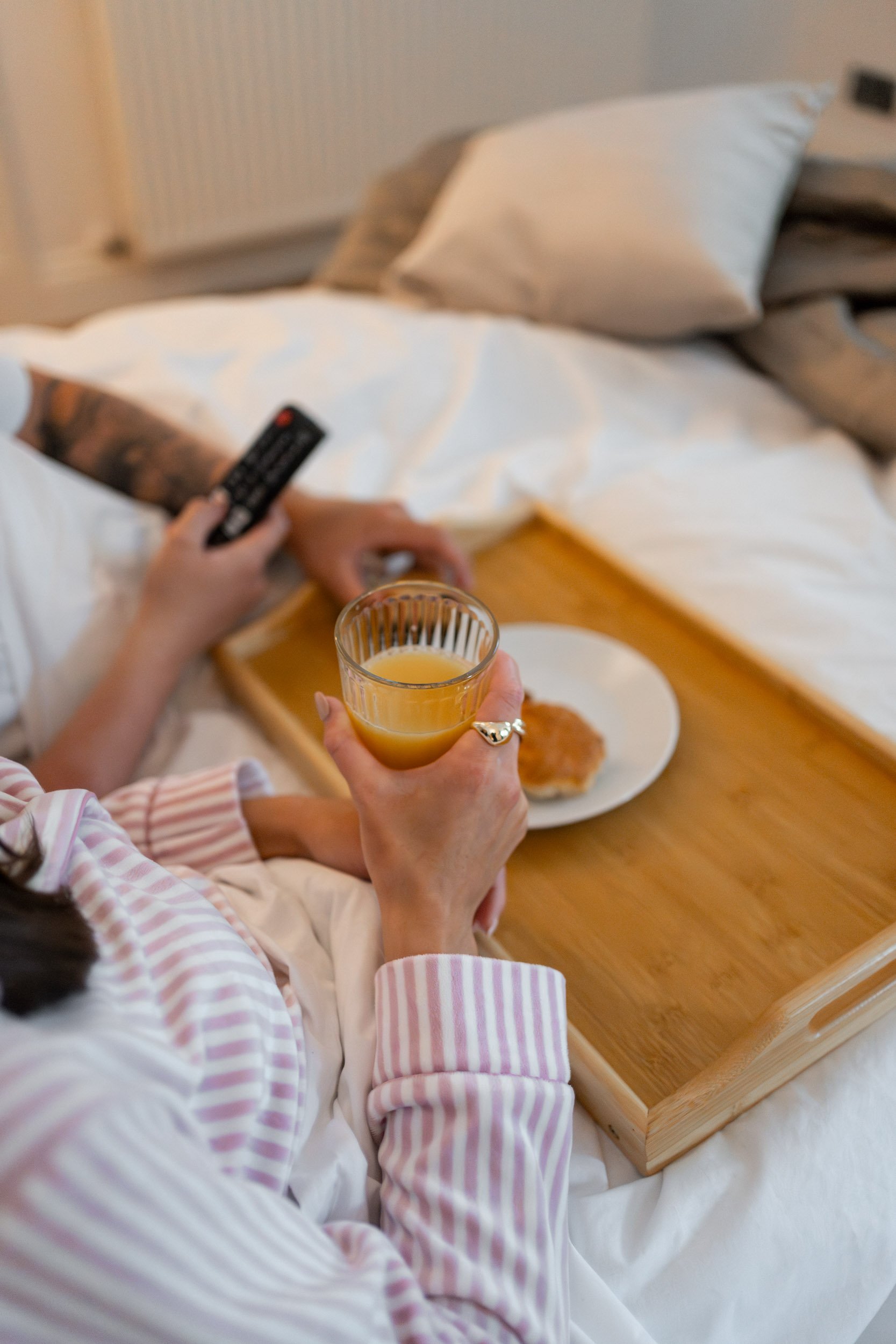 Person in striped pajamas holding a glass of orange juice, with a breakfast plate on a wooden tray, sitting on a bed with a tray and remote control in hand.
