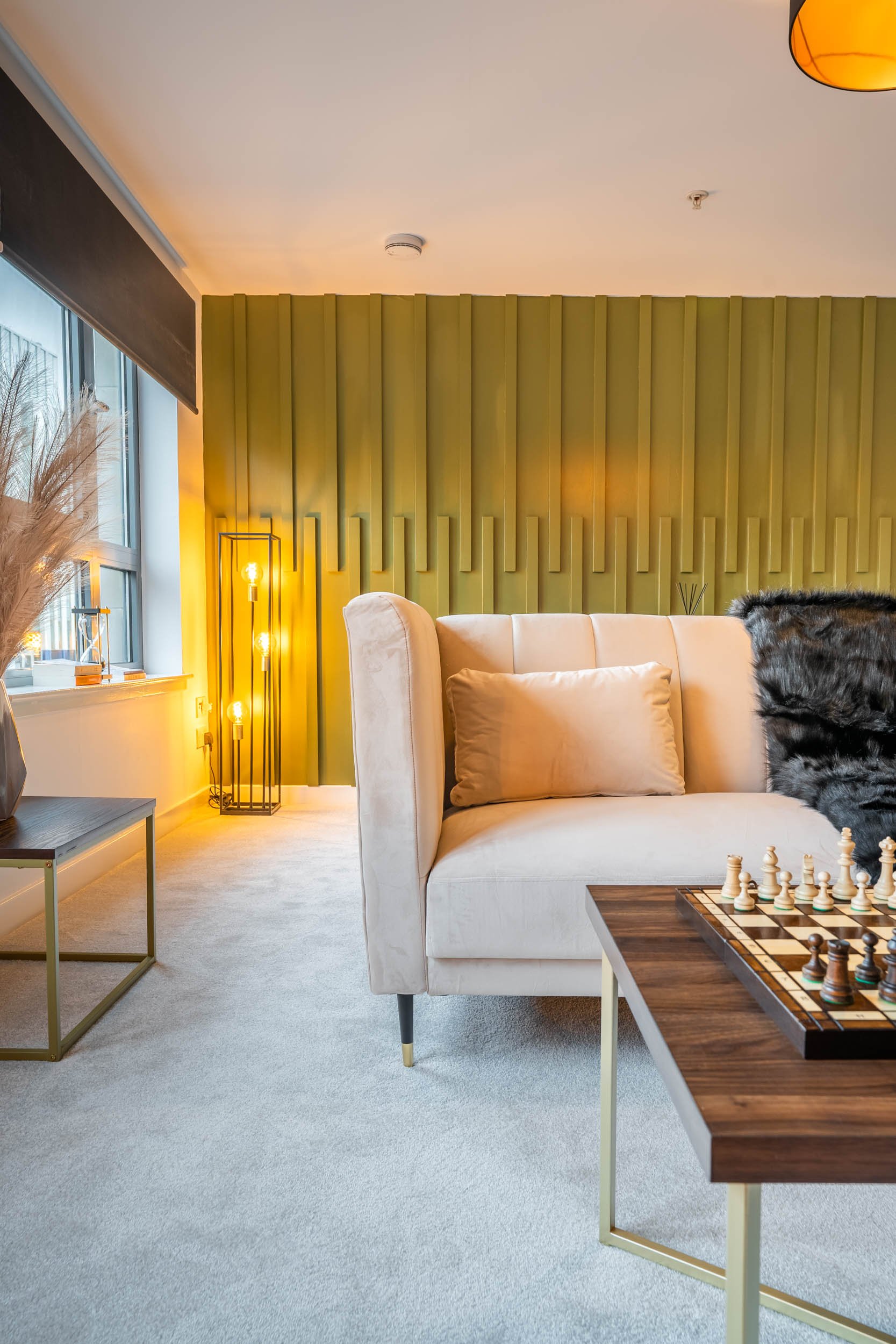 Modern living room with beige sofa, decorative pillows, chessboard on a wooden coffee table, green textured accent wall, tall black and gold floor lamp, window with blinds, beige carpet, and decorative pampas grass on ledge.