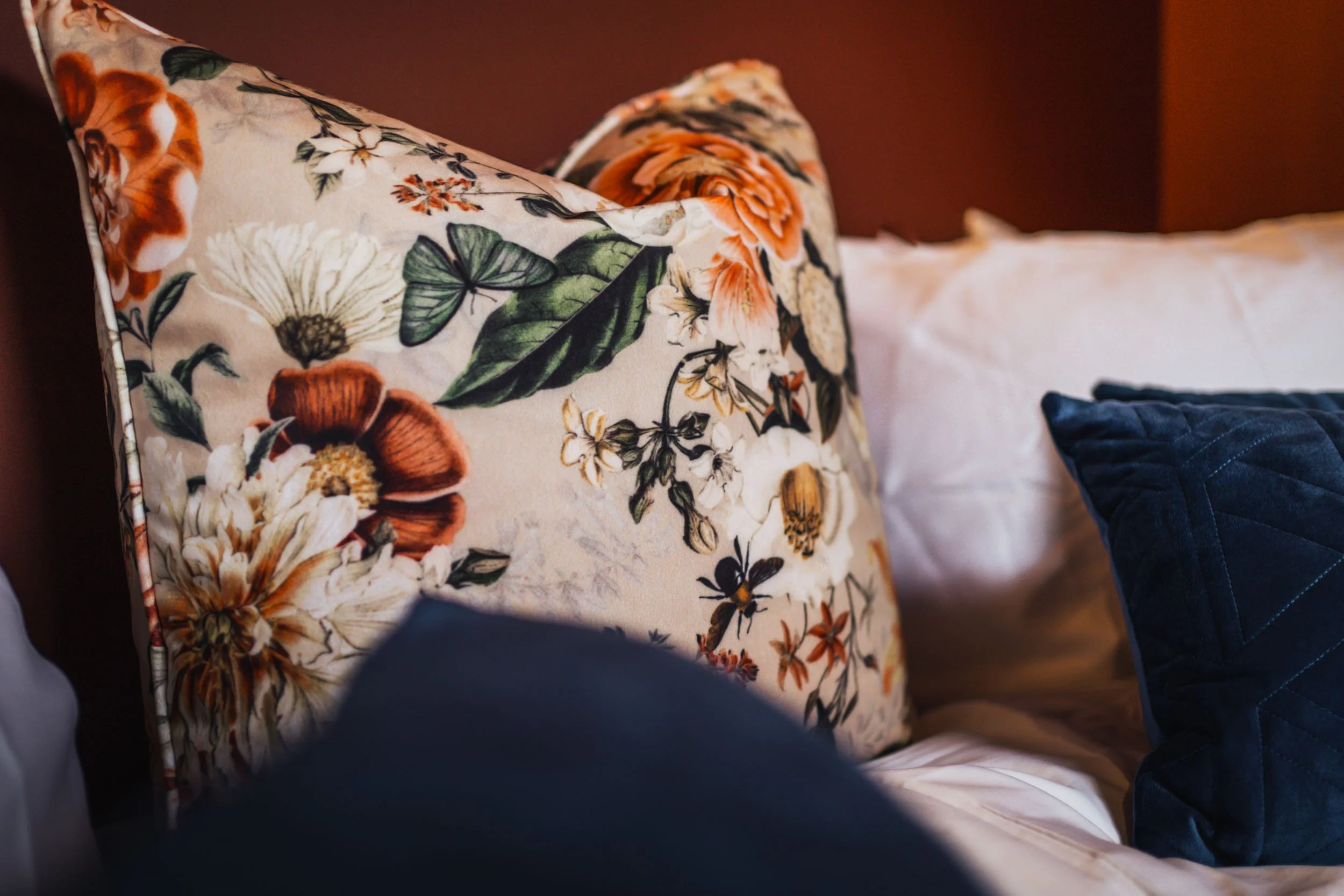 Decorative pillows on a bed, including a large floral-patterned cushion with orange, white, and green flowers, white pillow, and a navy blue quilted pillow.