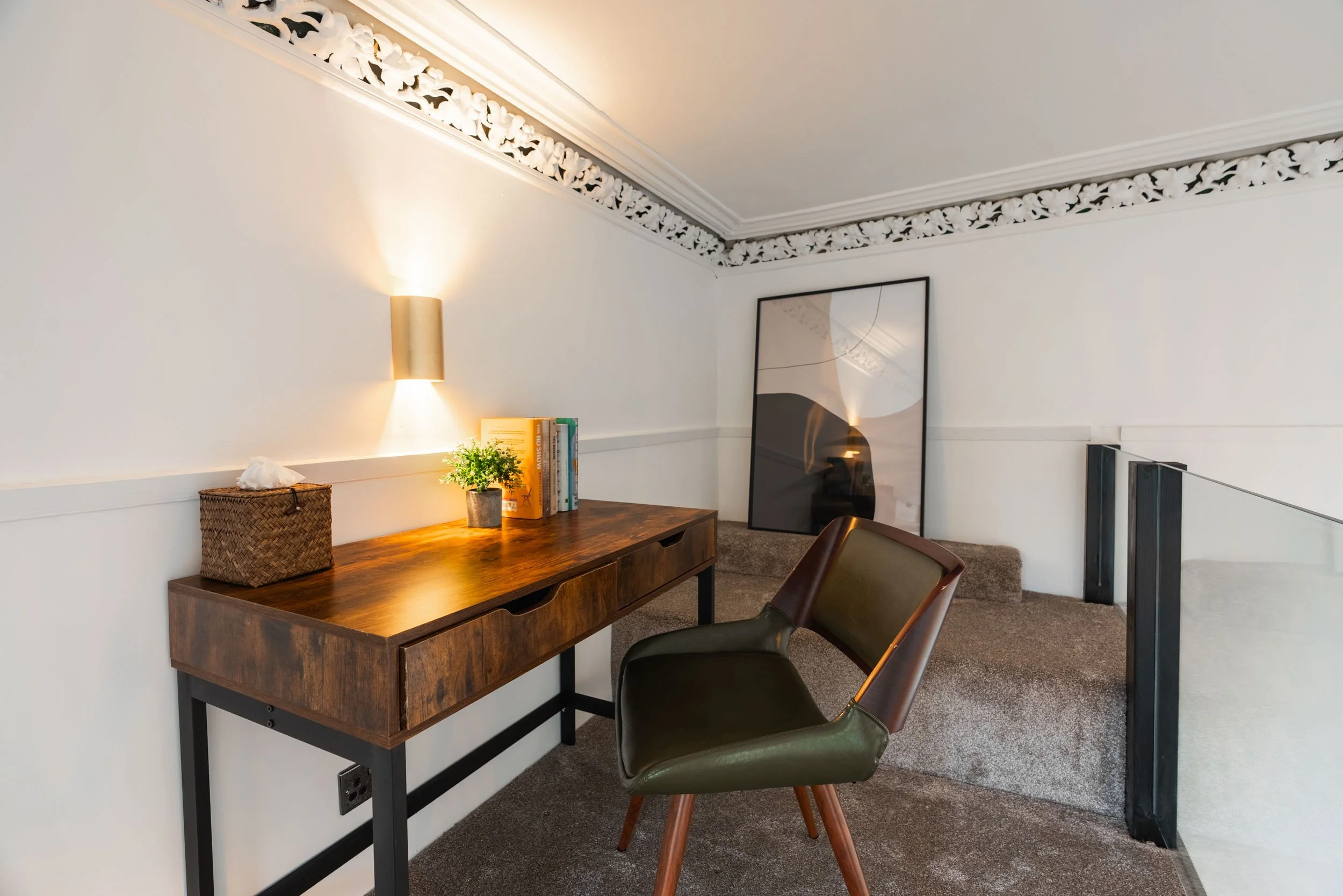 Home office with a wooden desk, green and brown chair, bookshelf, framed artwork, and a decorative wall trim.