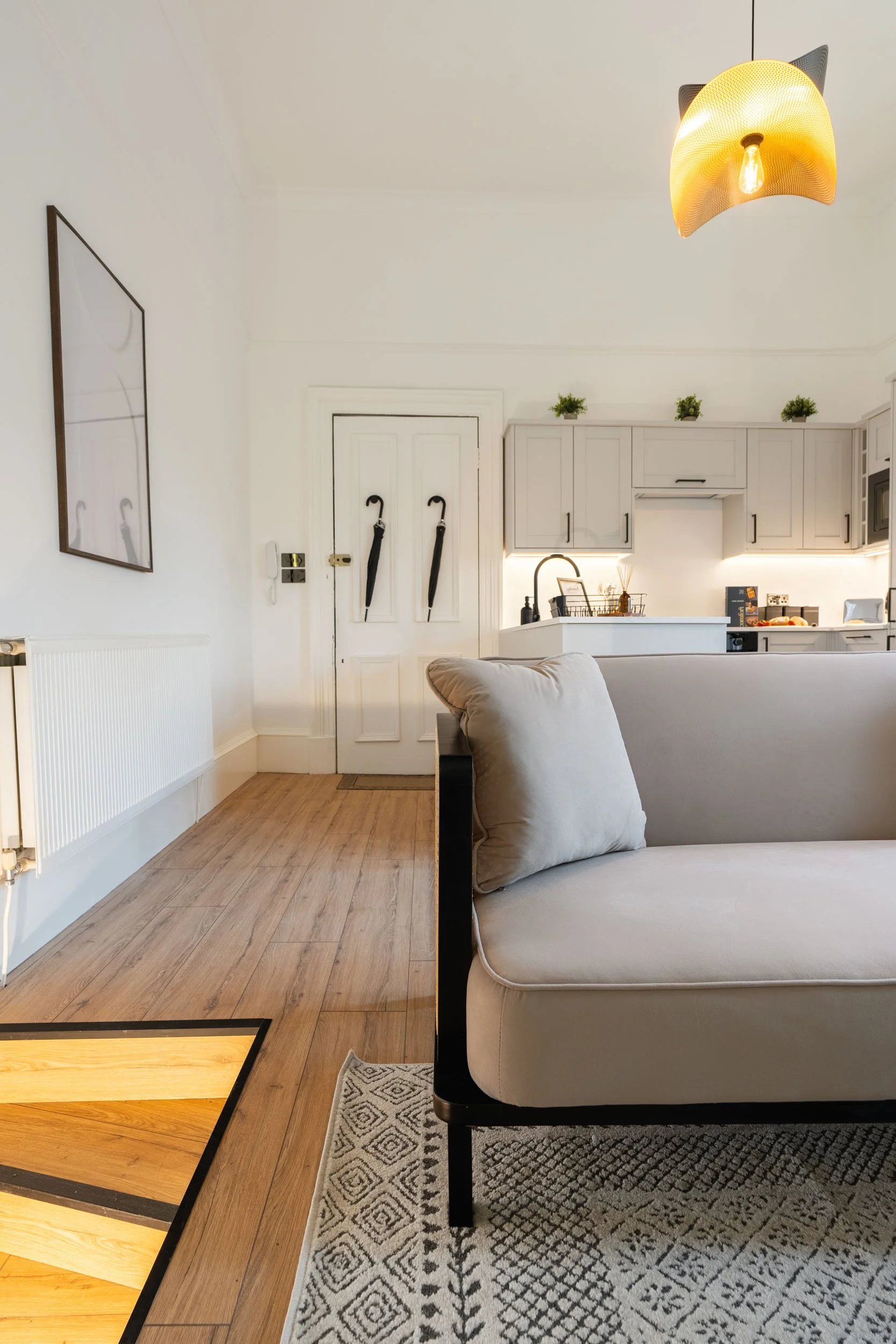 Interior of a modern, minimalist apartment with a white sofa, wooden flooring, a kitchen in the background, and closed white doors with two umbrellas hanging on them.